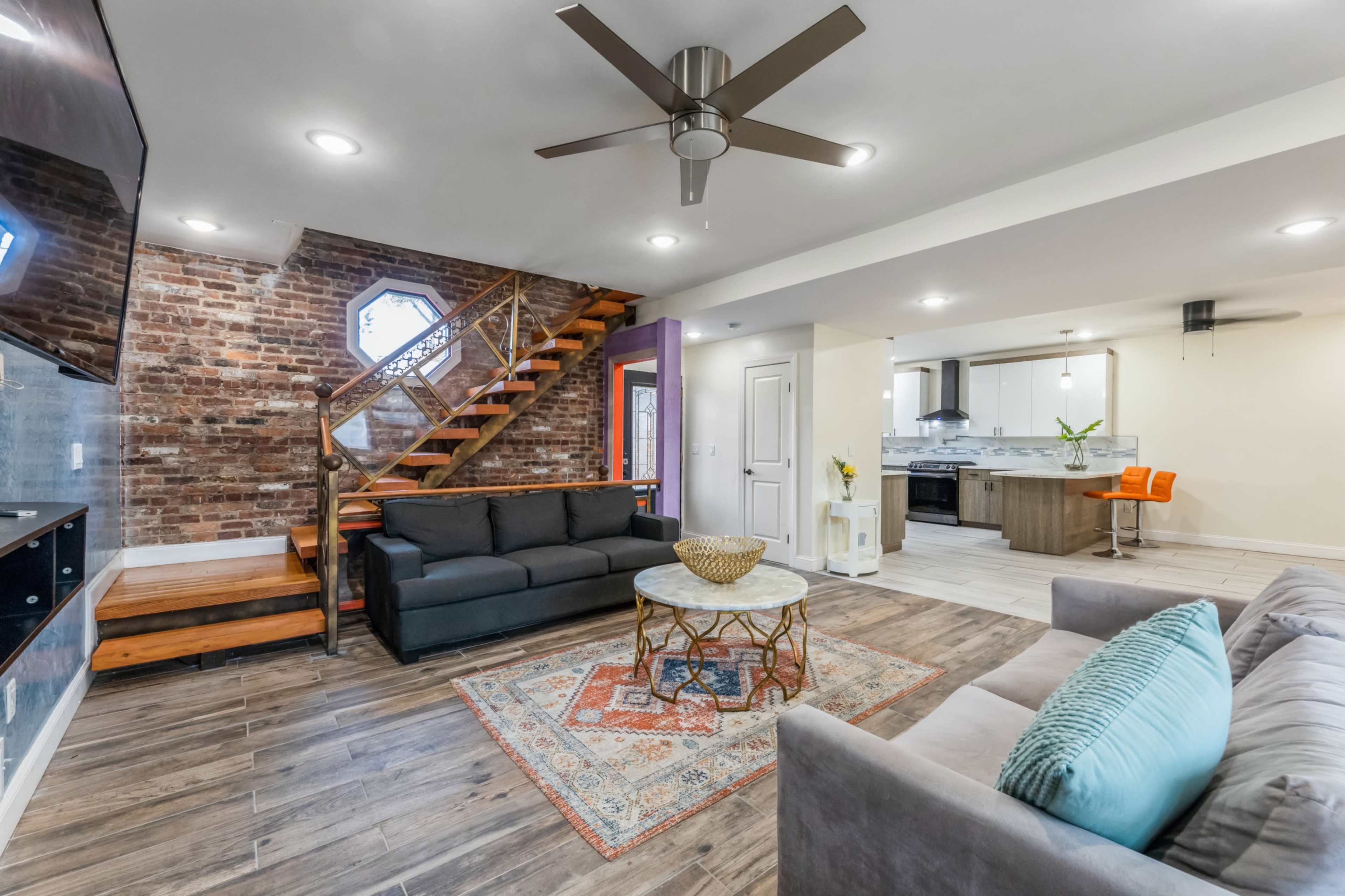 A modern living room with a brick wall, two sofas, a round coffee table on an area rug, and a staircase leading to an upper level.