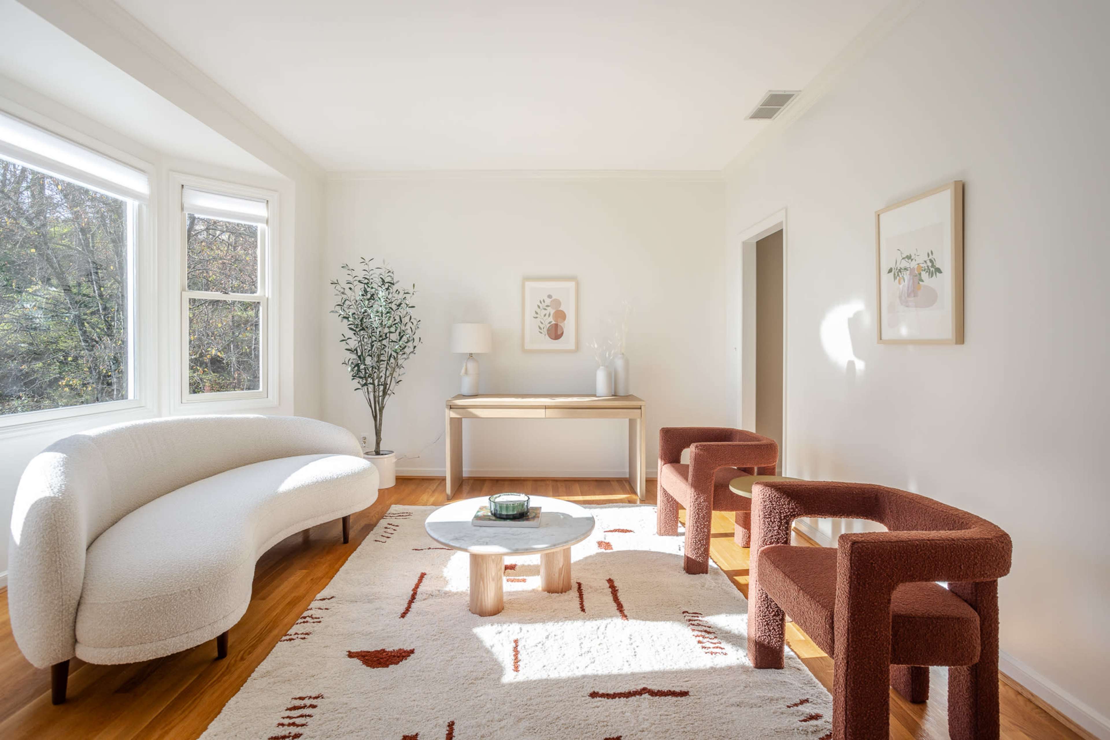 A bright, minimalist living room features a curved white sofa, two red upholstered chairs, a round coffee table, and a console table against a light-colored wall.