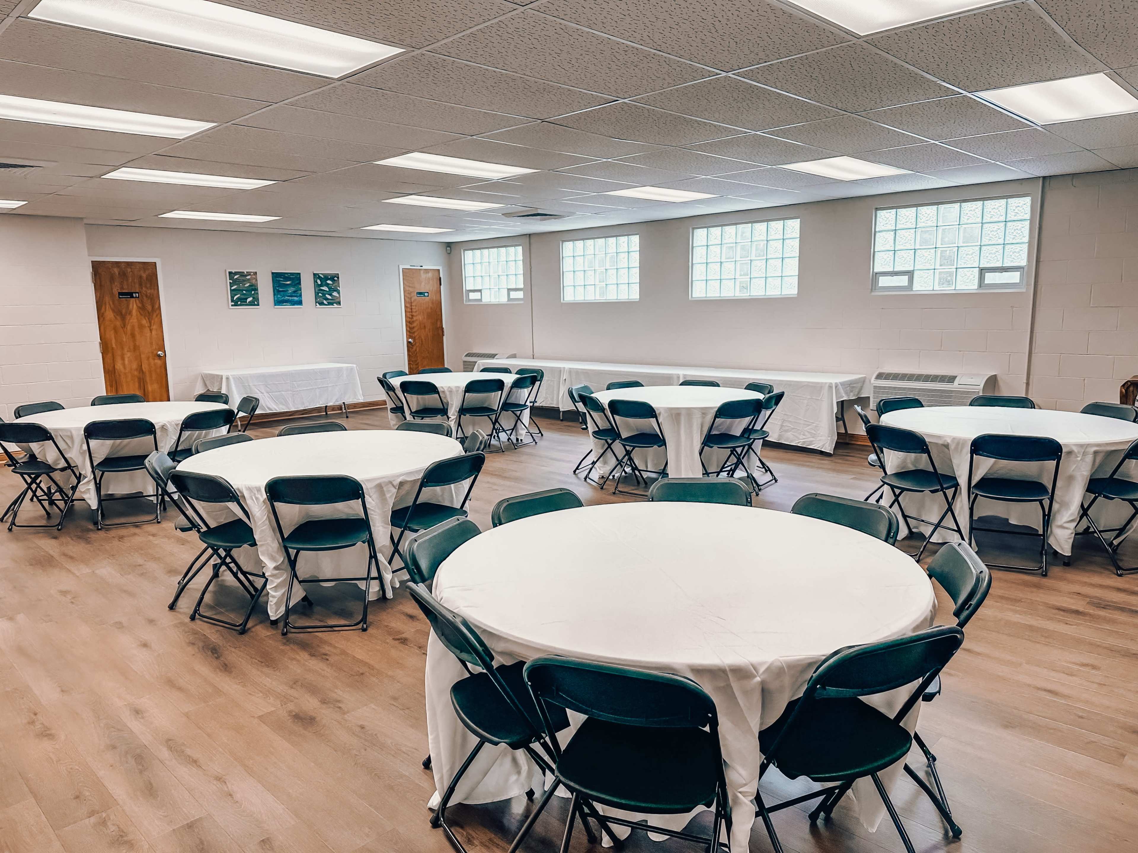 A room set up for an event, featuring several round tables covered with white tablecloths and green chairs around them.