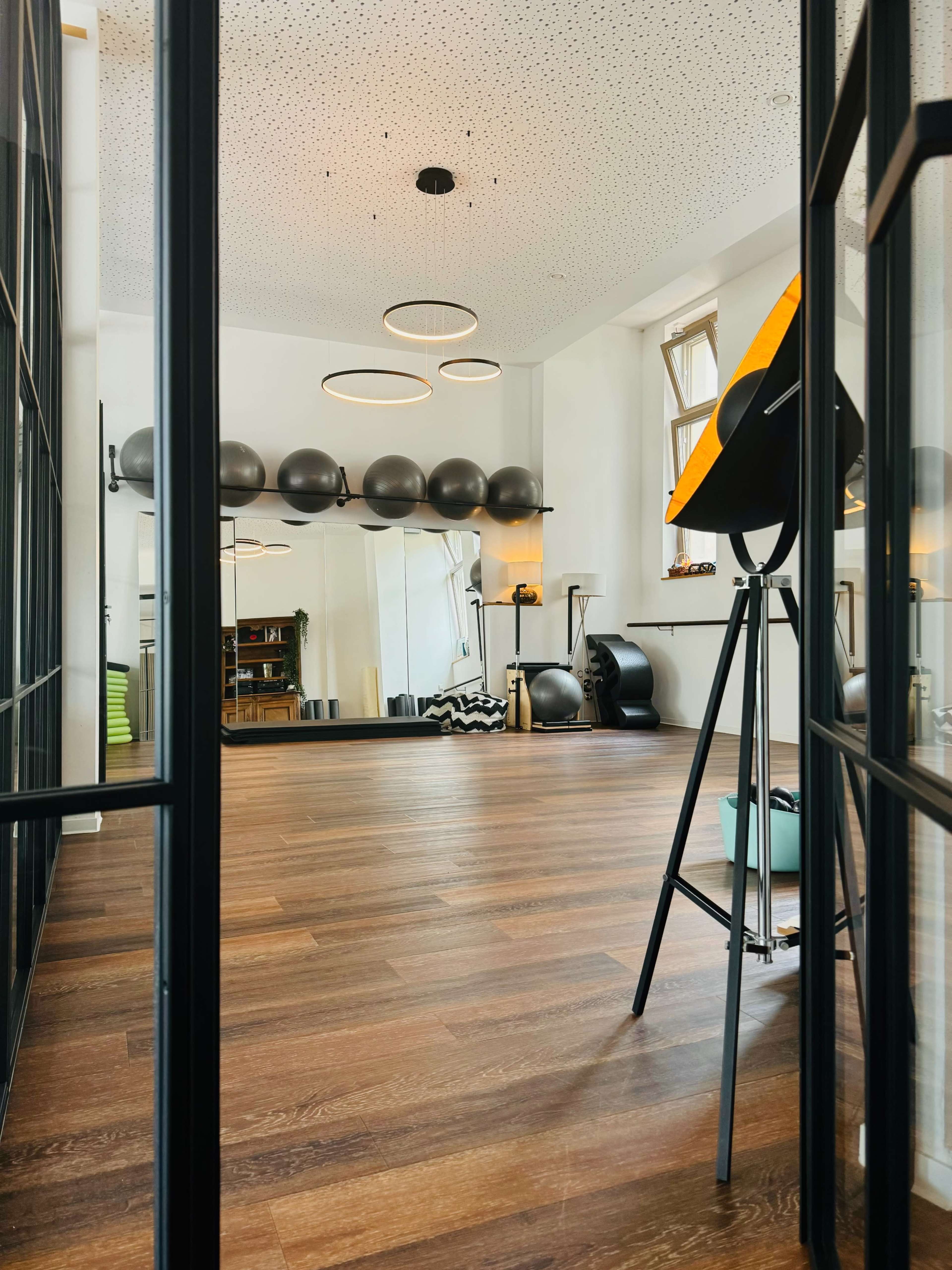The image shows a spacious, modern fitness studio with wooden flooring, fitness equipment, and large mirrors.