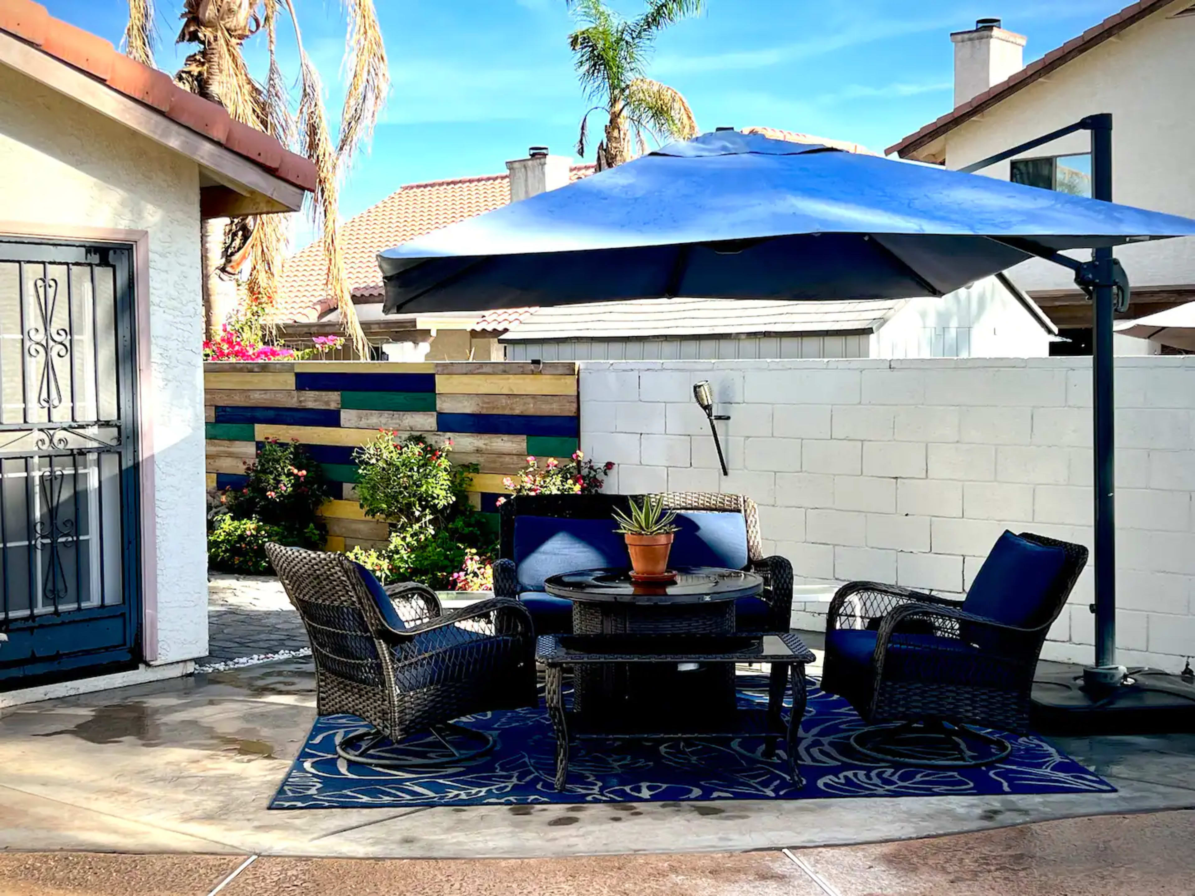 A shaded patio area features a round table surrounded by four chairs, with a potted plant on the table and colorful landscaping in the background.