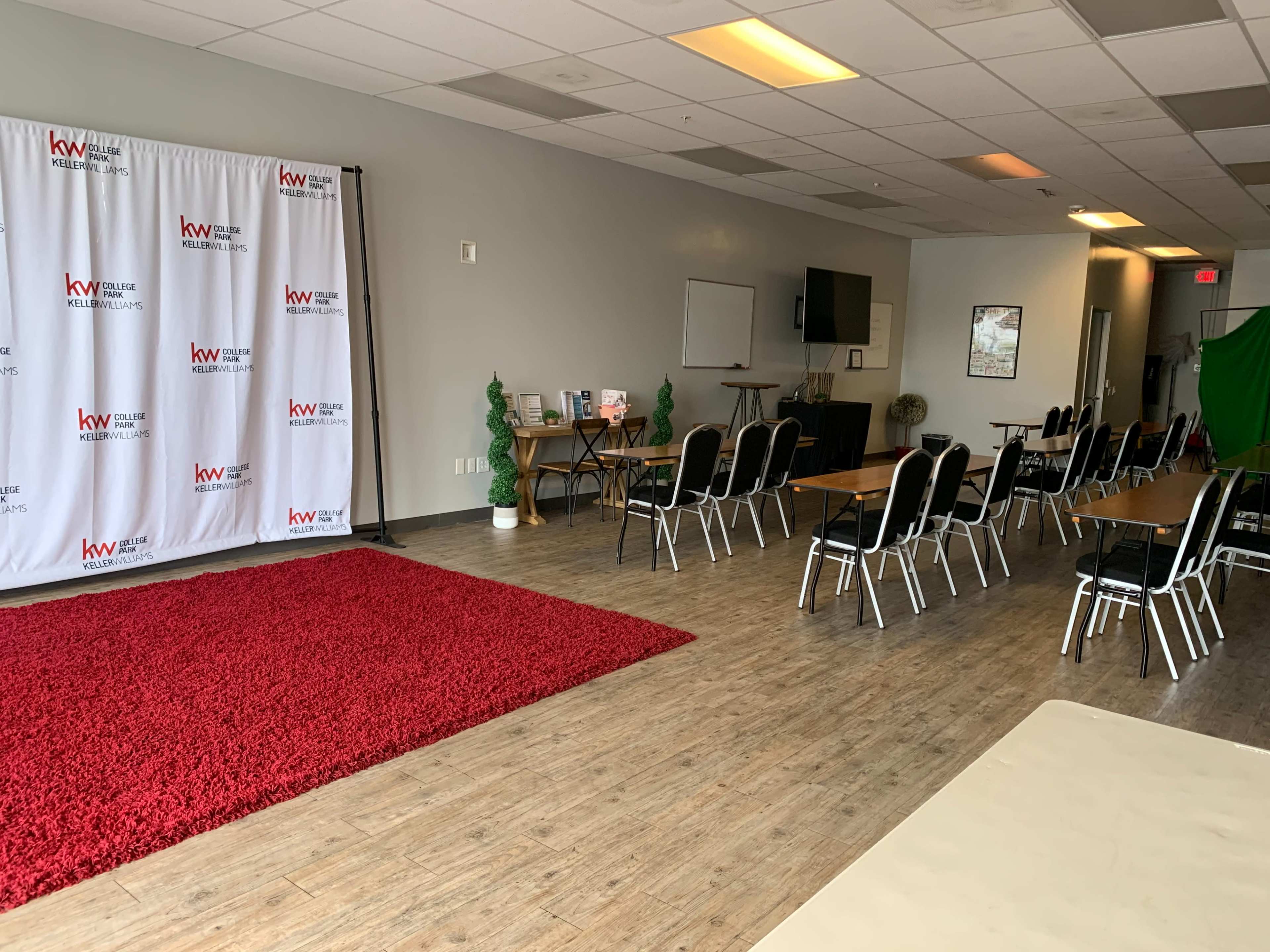 A meeting space with chairs arranged around tables, a red area rug, and a backdrop featuring a logo.