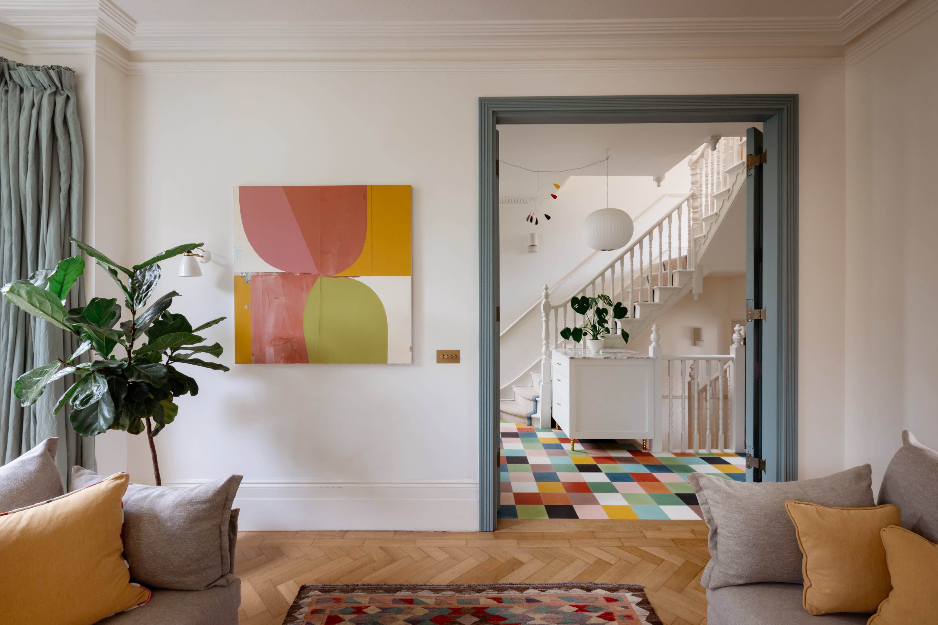 A modern living room features a large abstract painting, a potted plant, and comfortable sofas, with a view of a brightly colored hallway and staircase through an open door.