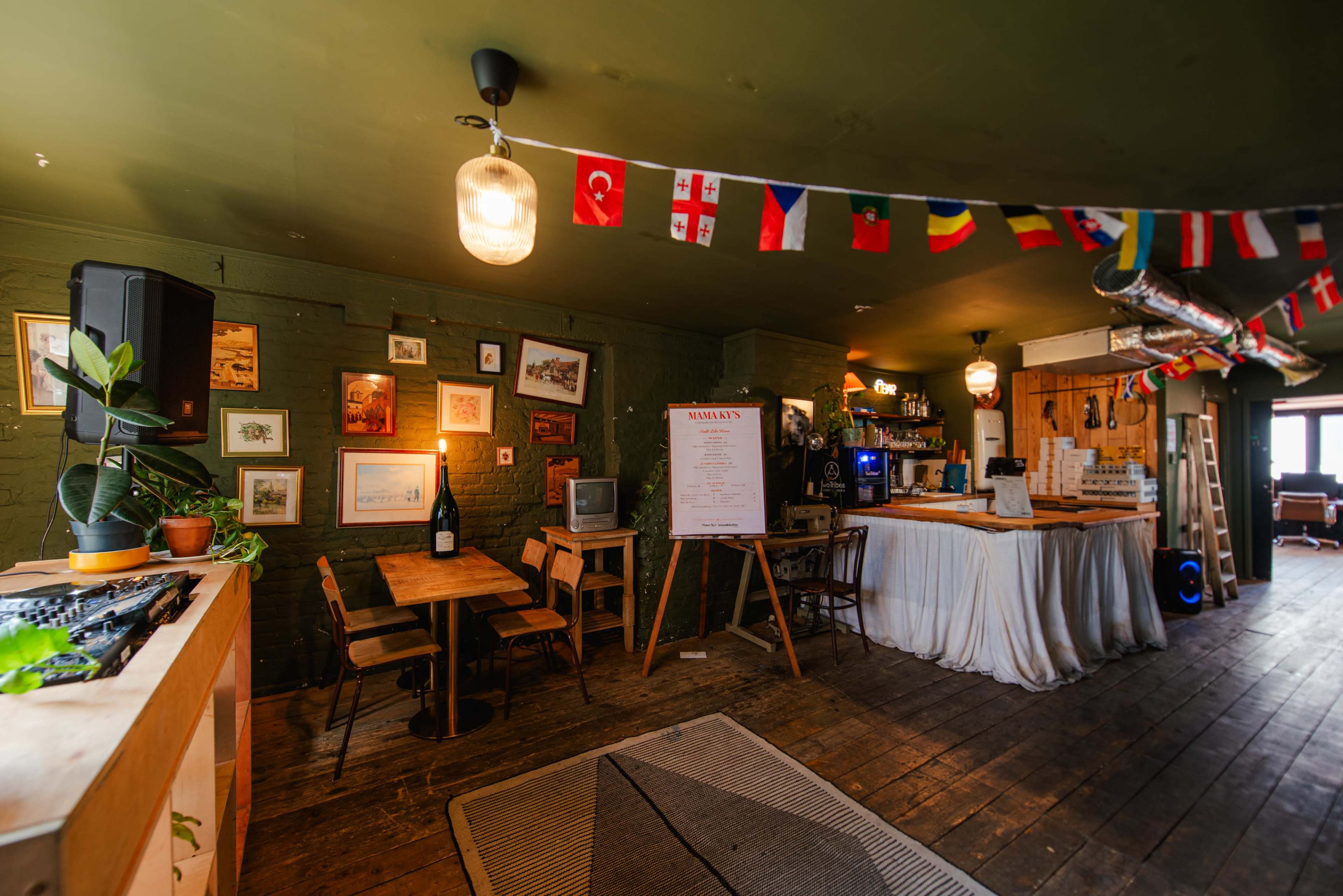 The image shows a cozy café interior with wooden flooring, hanging flags, a bar area, and various decorations on the walls.