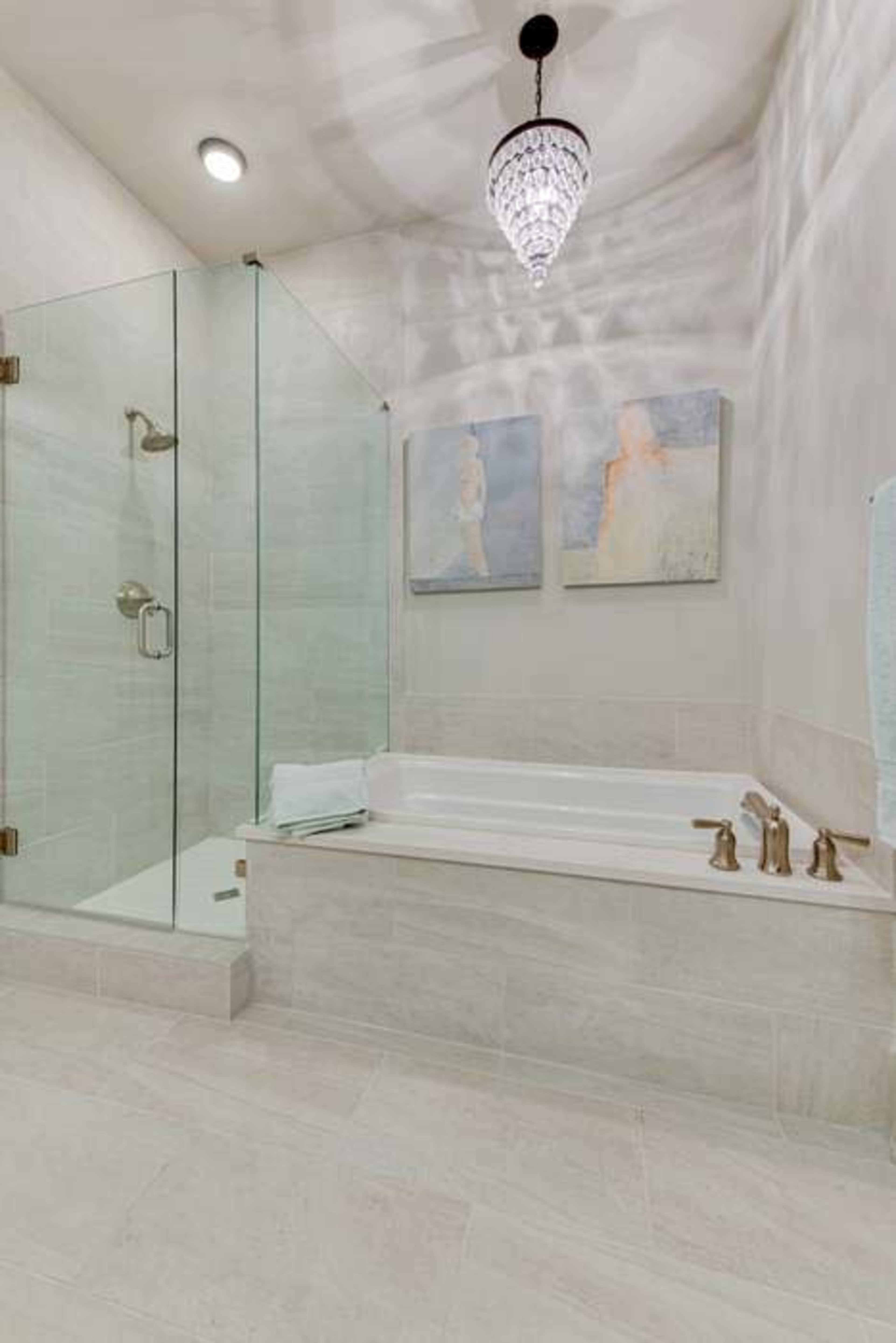 The image shows a modern bathroom featuring a glass shower enclosure, a freestanding bathtub, and wall art above the tub, illuminated by a decorative chandelier.