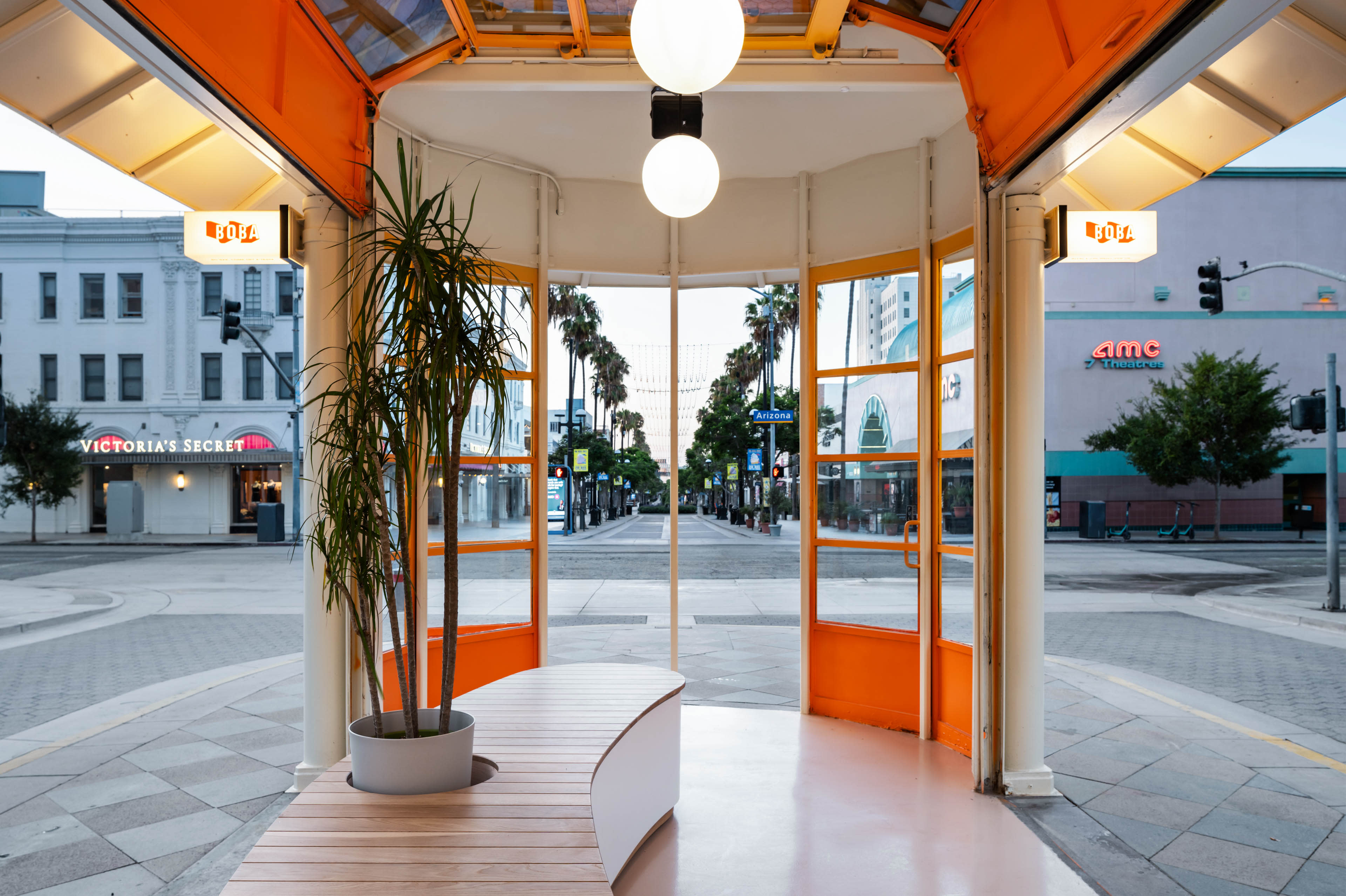 The image shows a modern pavilion with an orange and white interior, featuring a potted plant, overlooking a deserted street lined with palm trees and storefronts.