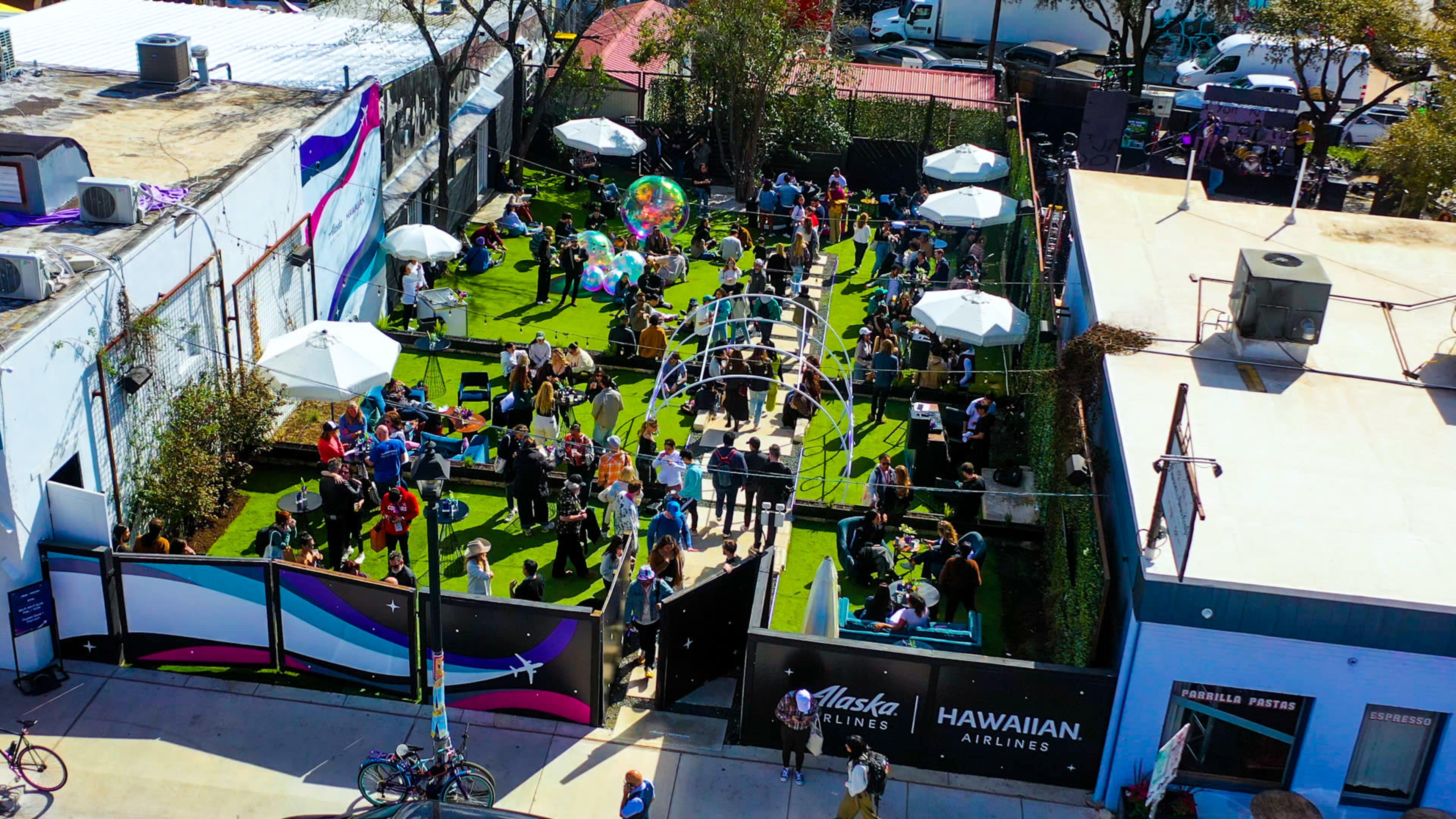 A crowded outdoor event space features guests socializing under white umbrellas on green grass, surrounded by food vendors and decorations.