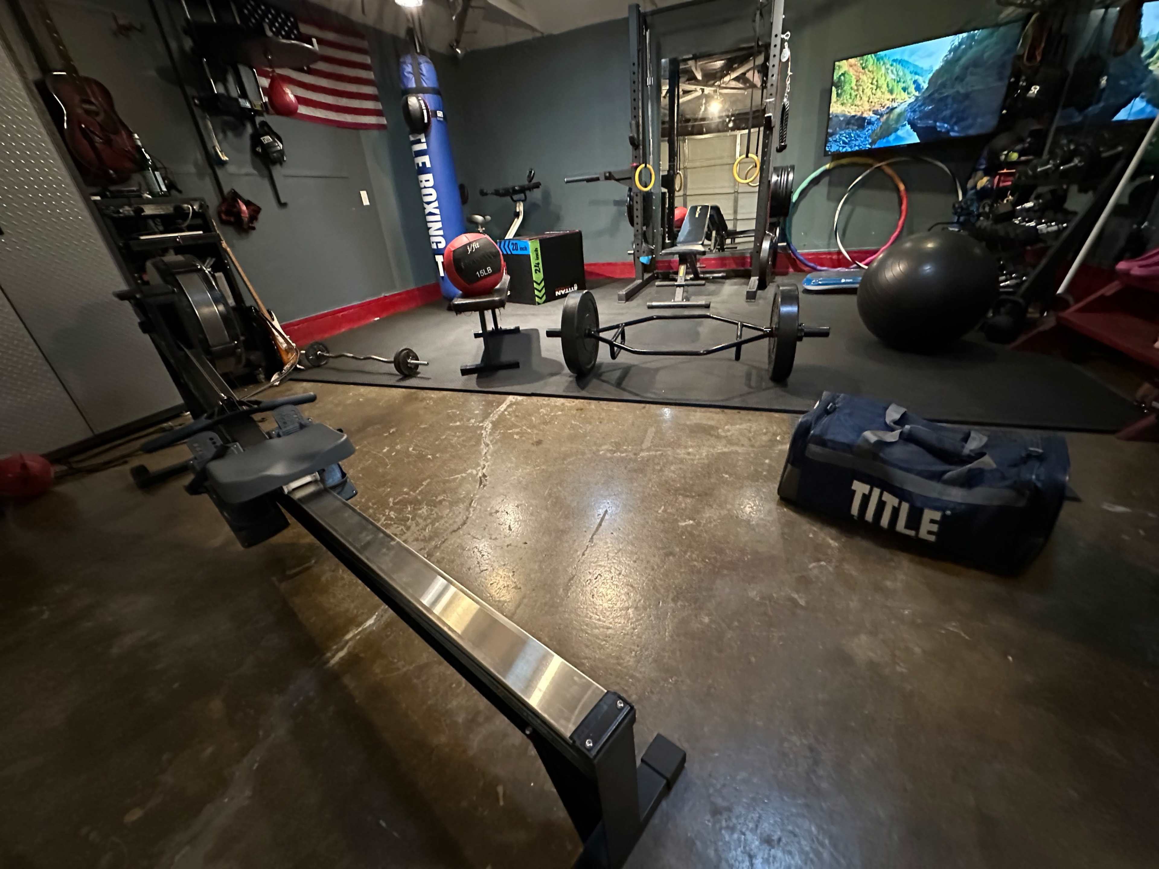 A home gym featuring various exercise equipment, including a rowing machine, weights, and a stability ball, arranged in a concrete-floored space with a screen displaying a nature scene.