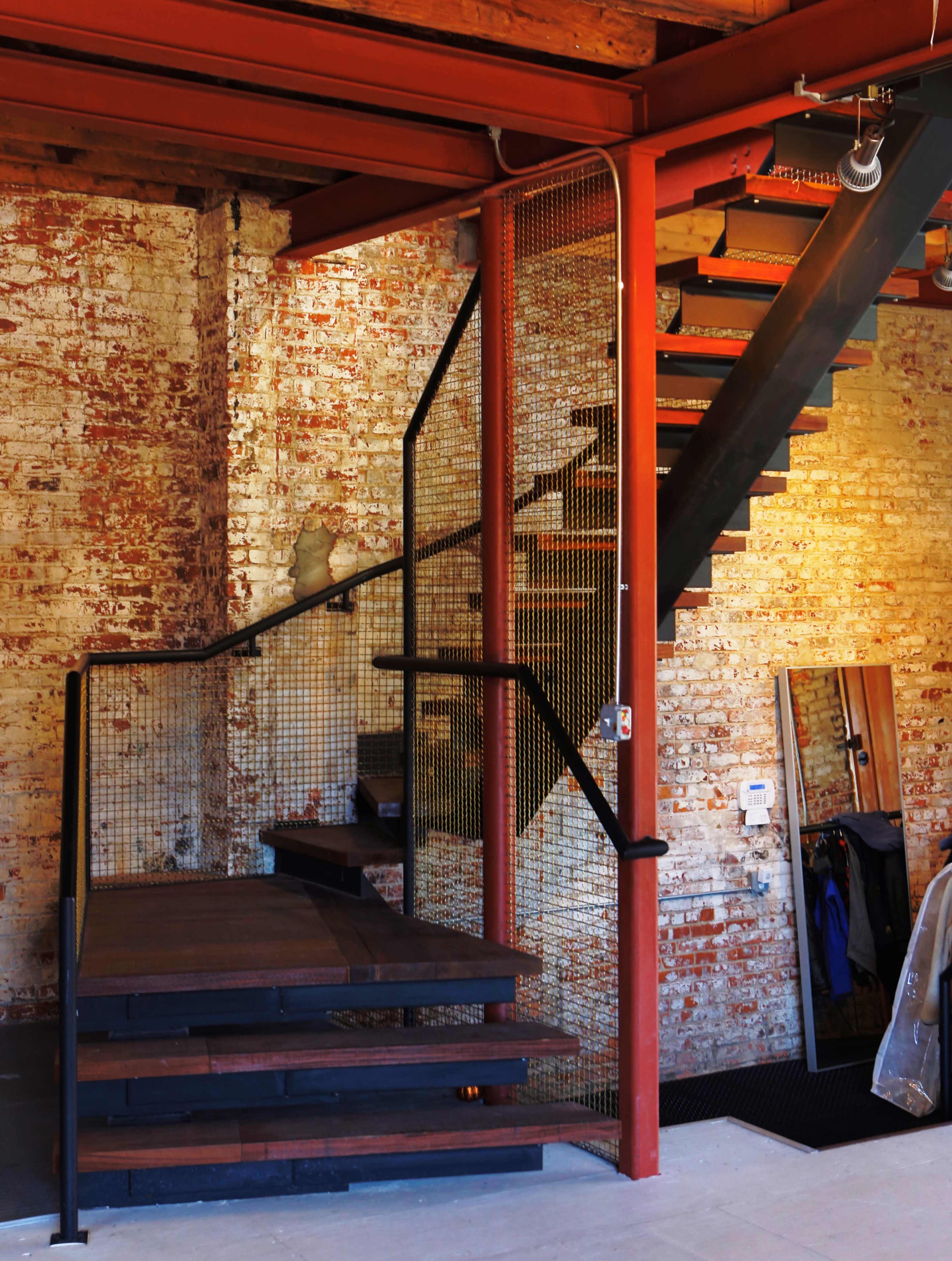 The image shows a modern staircase with wooden steps and a black metal railing, contrasting against a brick wall in a bright interior space.