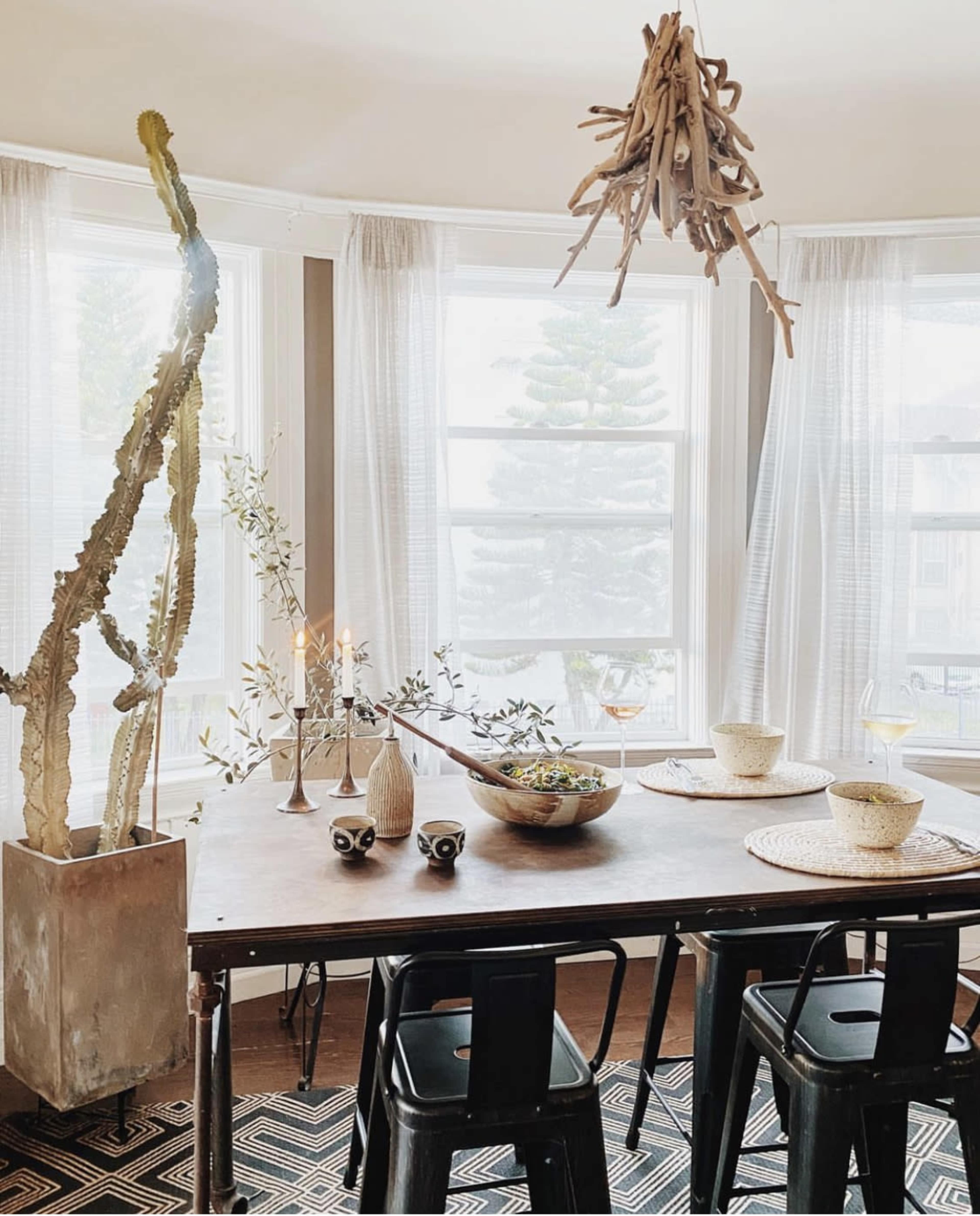 A dining table is set with a large bowl of salad, surrounded by four black chairs, in a sunlit room with large windows and a hanging decorative piece made of branches.