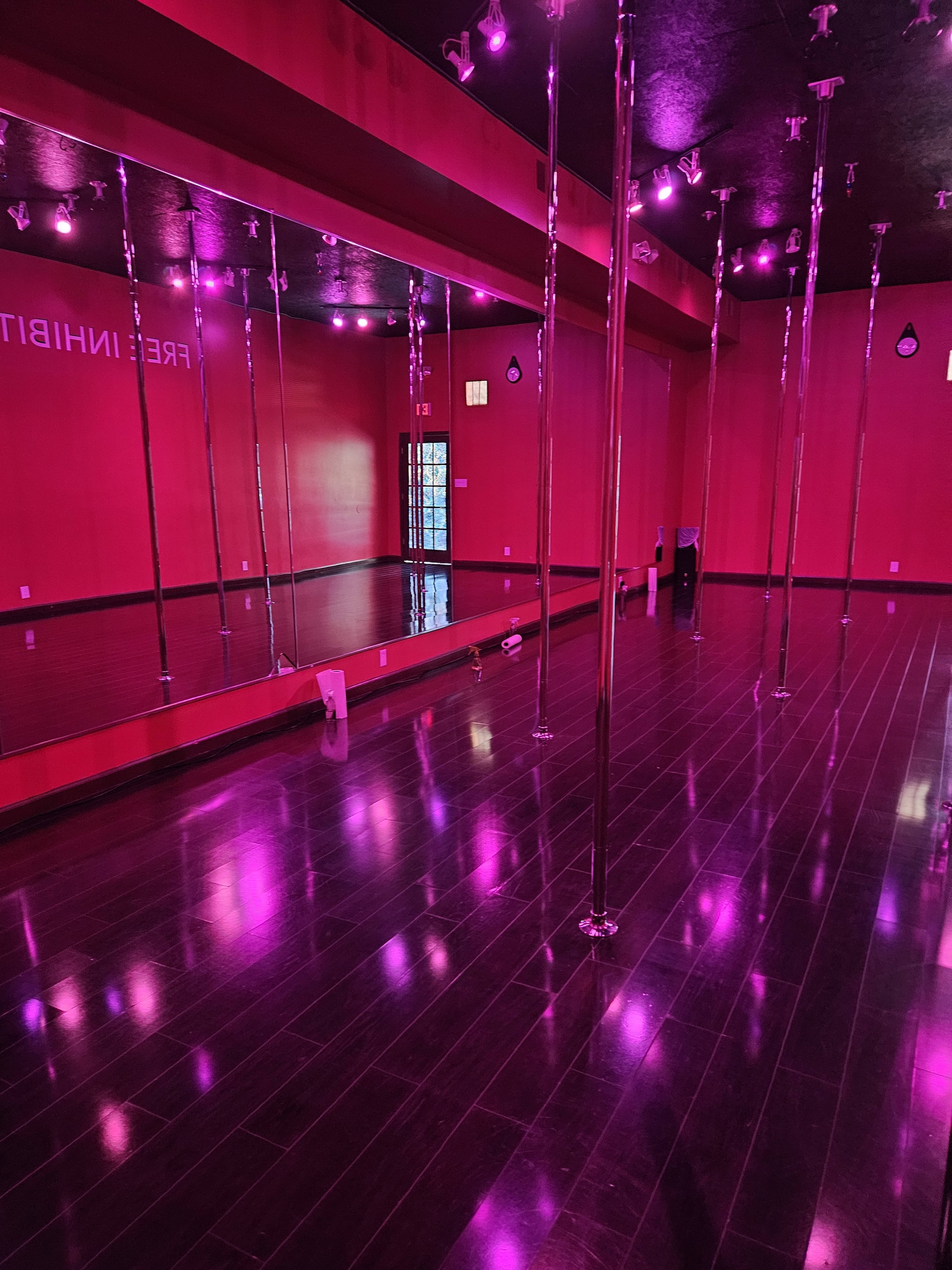 A brightly lit dance studio with pink walls, polished wooden floors, and several mirror-backed poles.