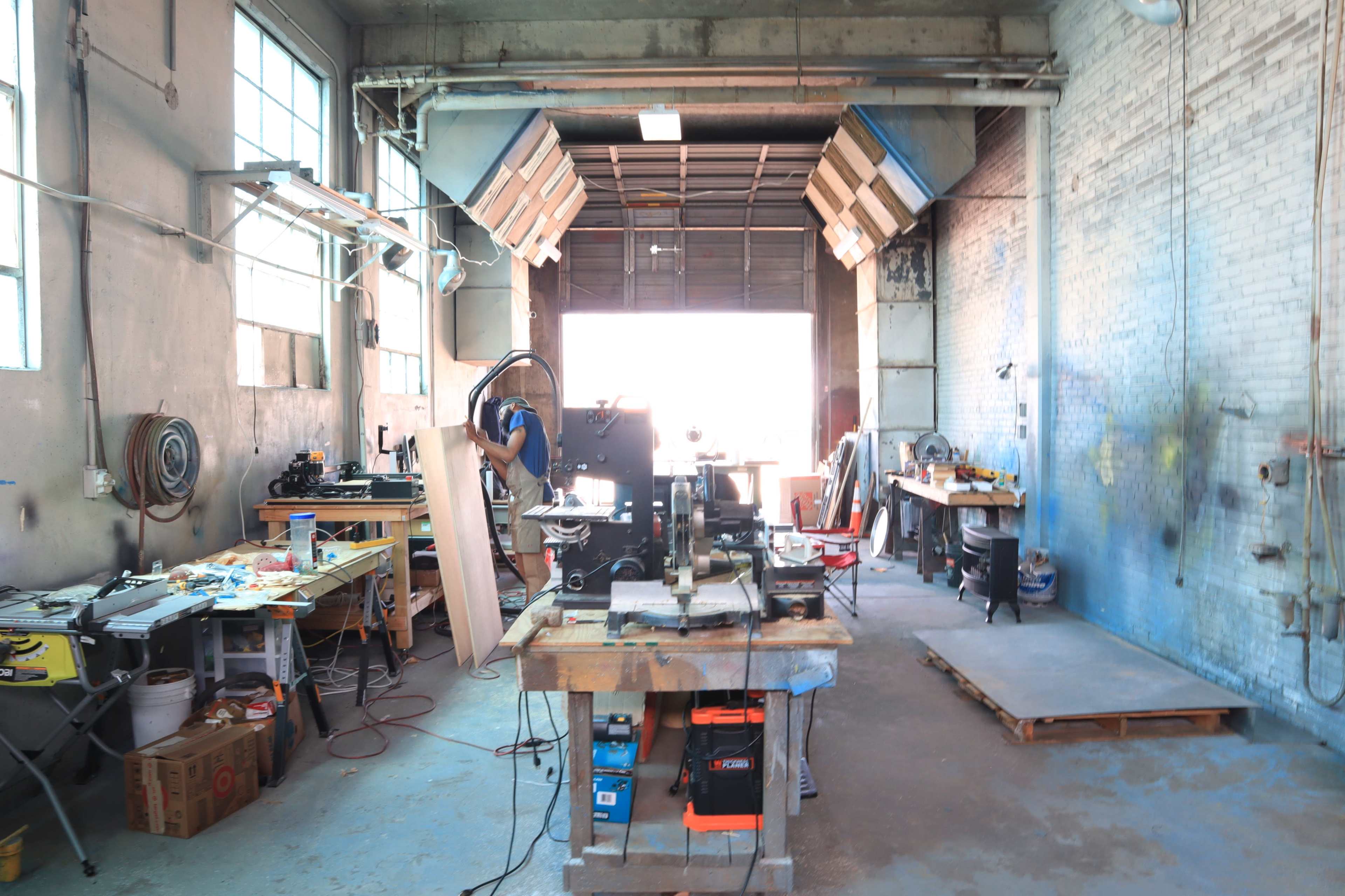 The image shows a workshop interior with various tools and equipment arranged around a central workspace, illuminated by natural light from a large open doorway.