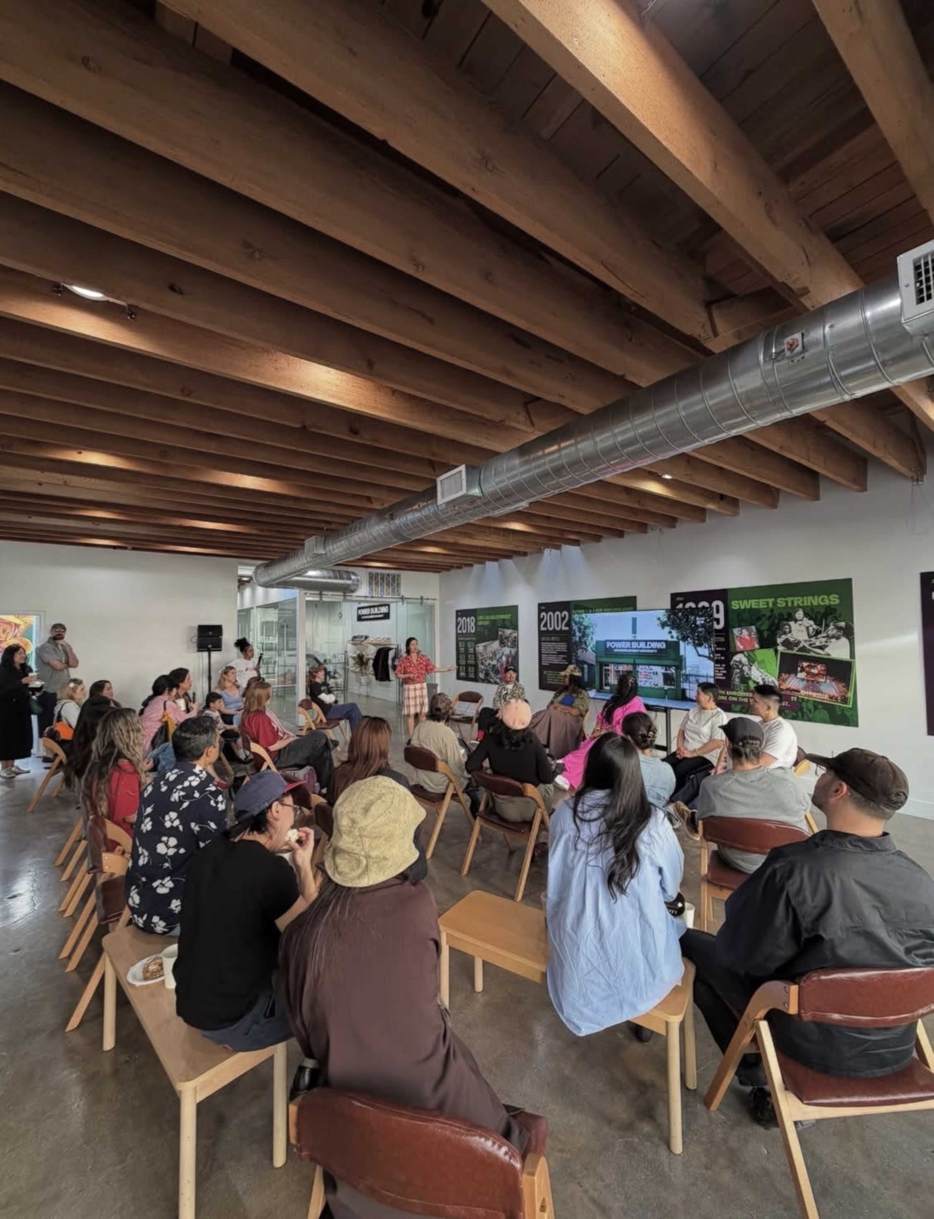 A group of people sits on wooden chairs in a spacious room with wooden beams, attending a presentation on a screen.