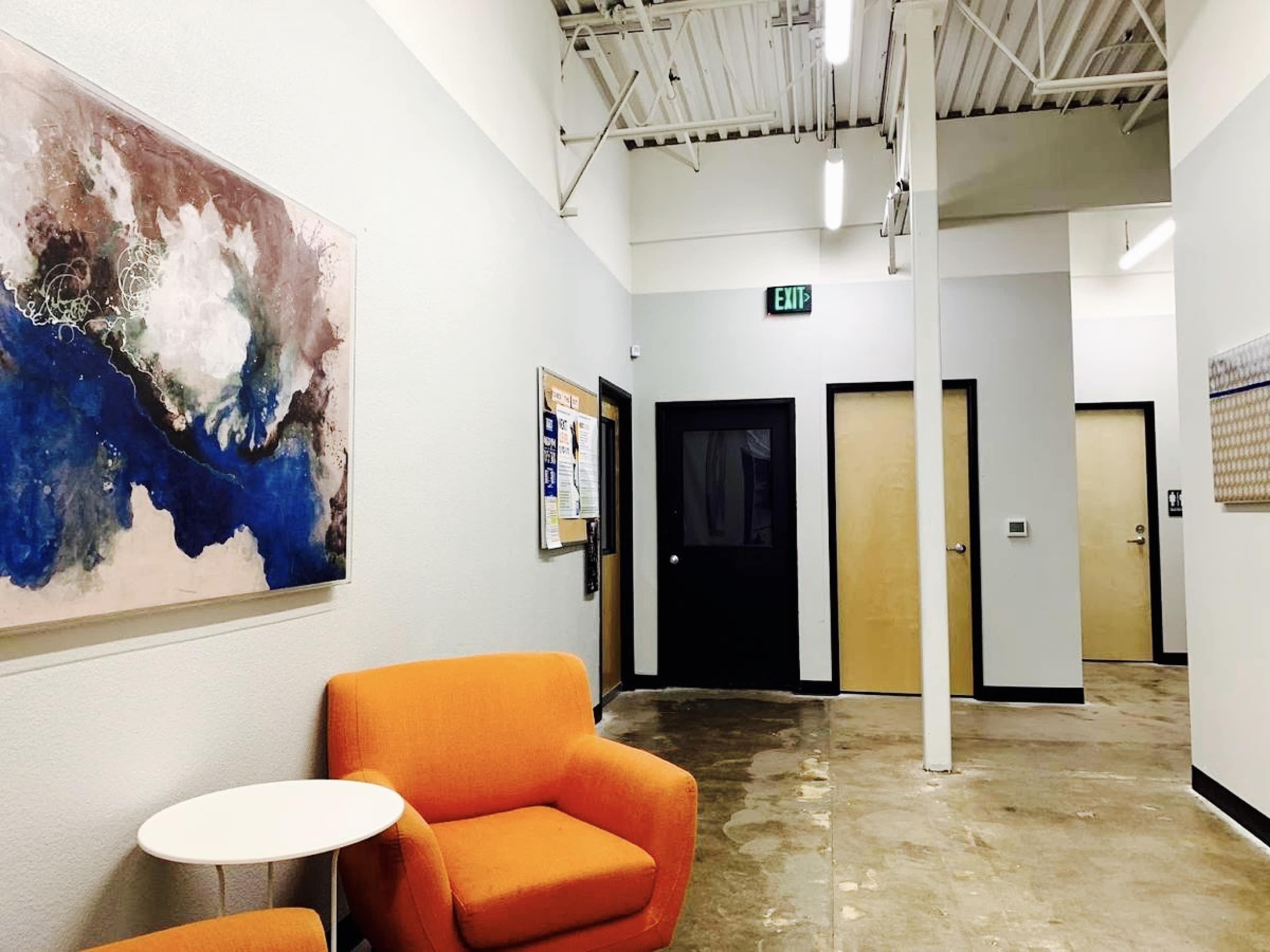The image shows a corridor with two orange chairs and a small round table, alongside walls featuring art and a bulletin board.