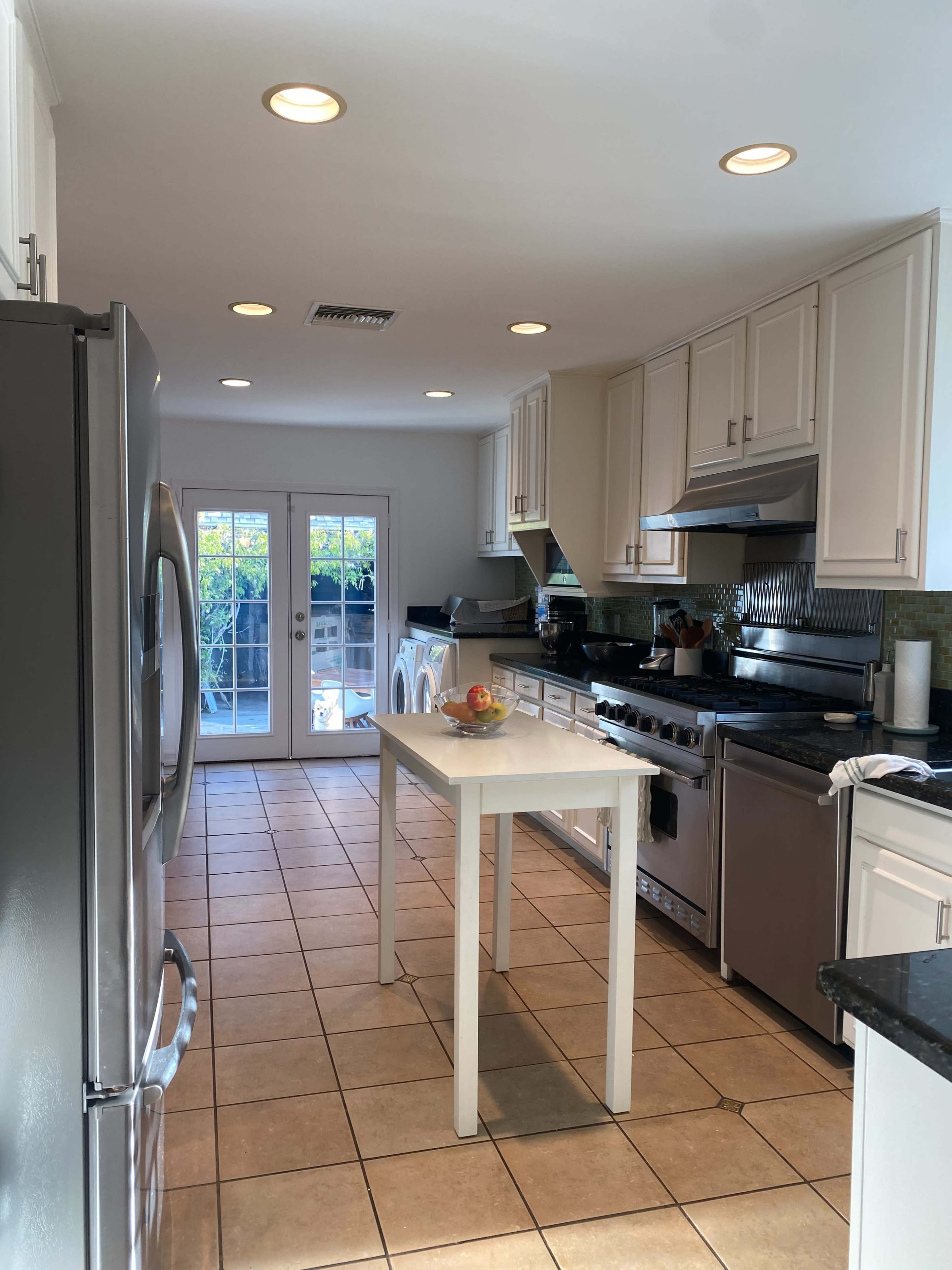 The image shows a modern kitchen featuring stainless steel appliances, a central white table, and a door leading to an outdoor area.