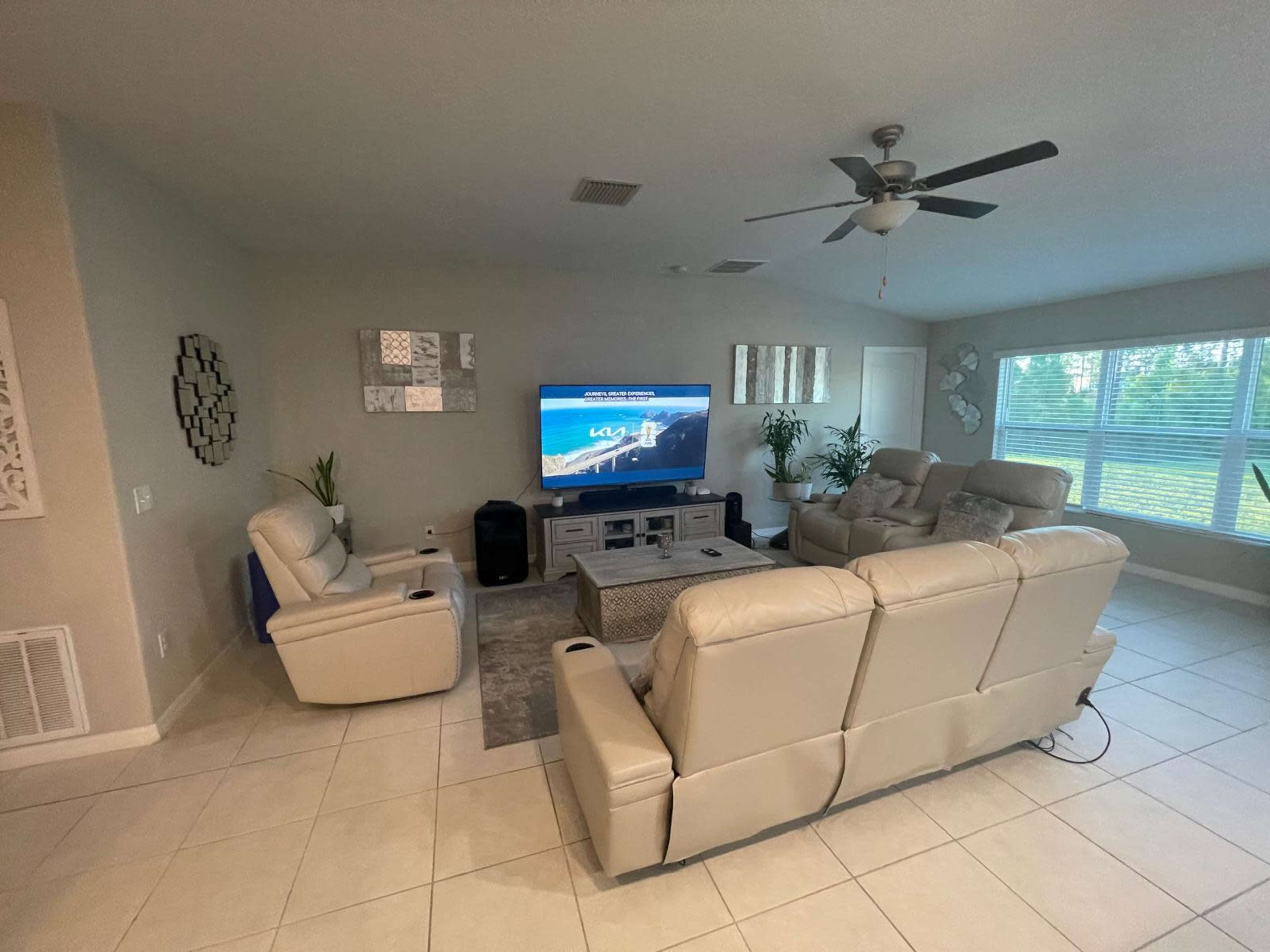 The living room features a light-colored seating arrangement facing a television, with large windows allowing natural light to enter.