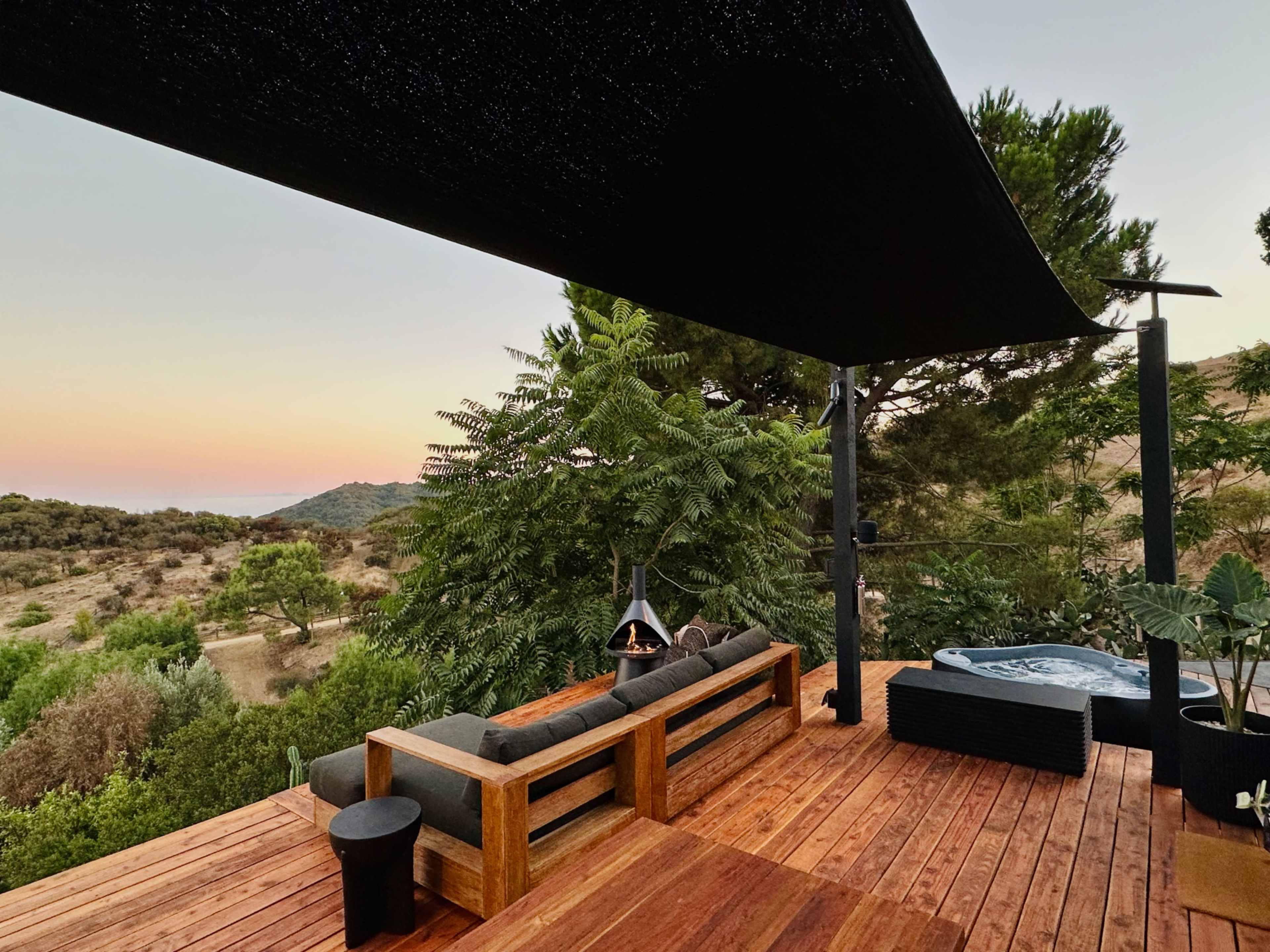 A wooden deck features a seating area and a hot tub, surrounded by trees and hills under a twilight sky.