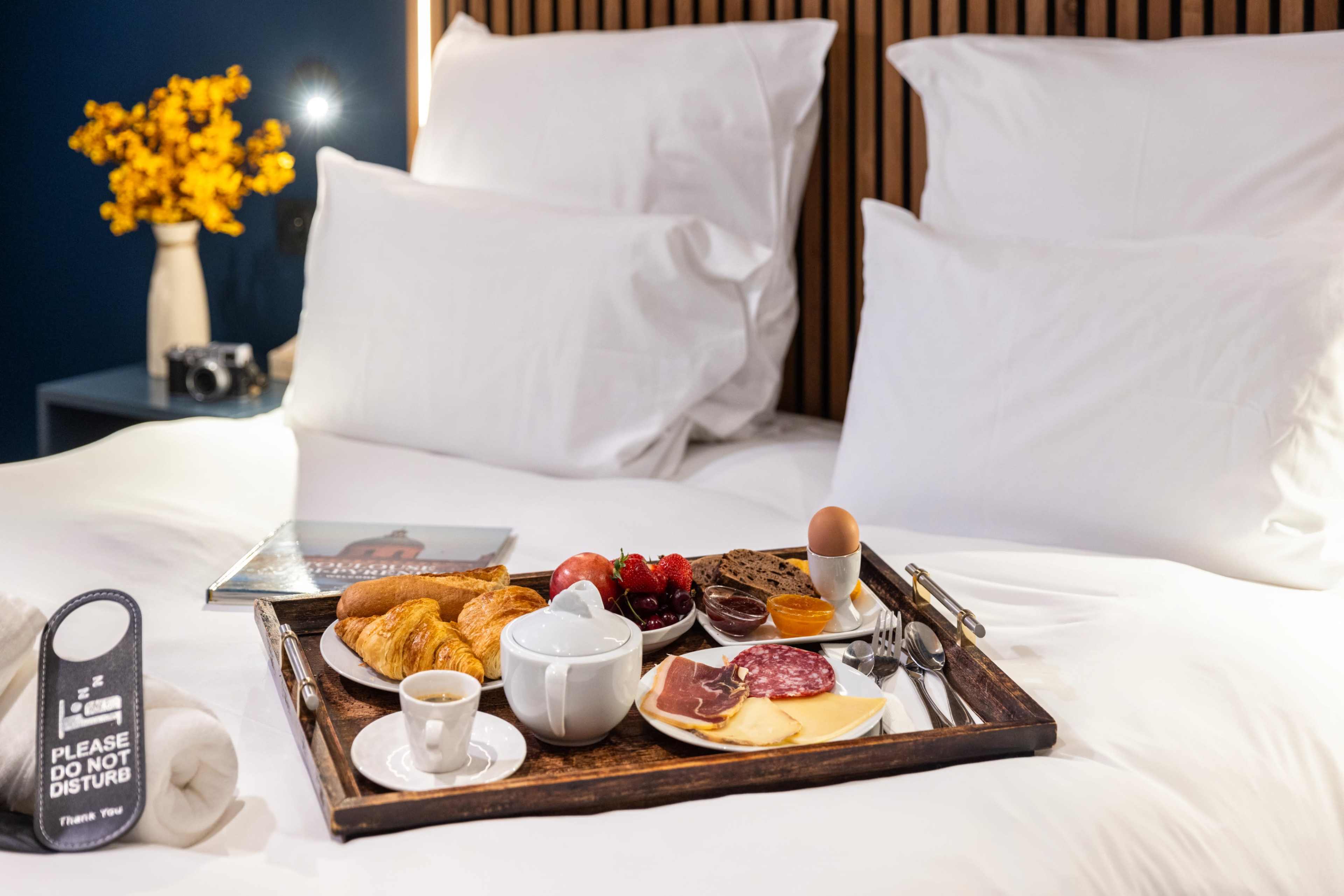 A breakfast tray with various foods, including croissants, cheese, meats, tomatoes, an egg, and coffee, is placed on a bed with white linens and fluffy pillows.