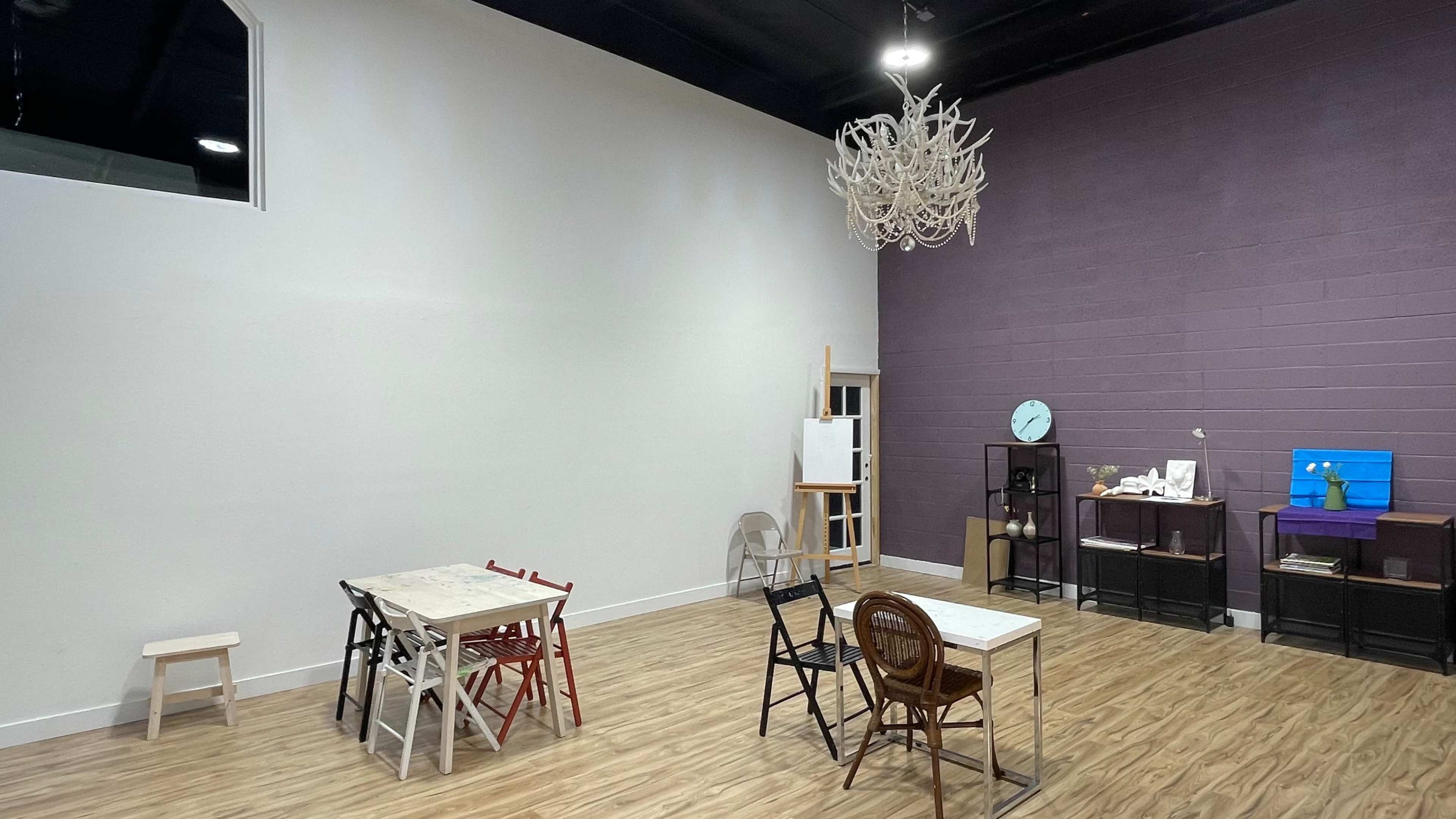 A spacious room with wooden flooring, featuring several tables and chairs along with a wall-mounted clock and a decorative chandelier.
