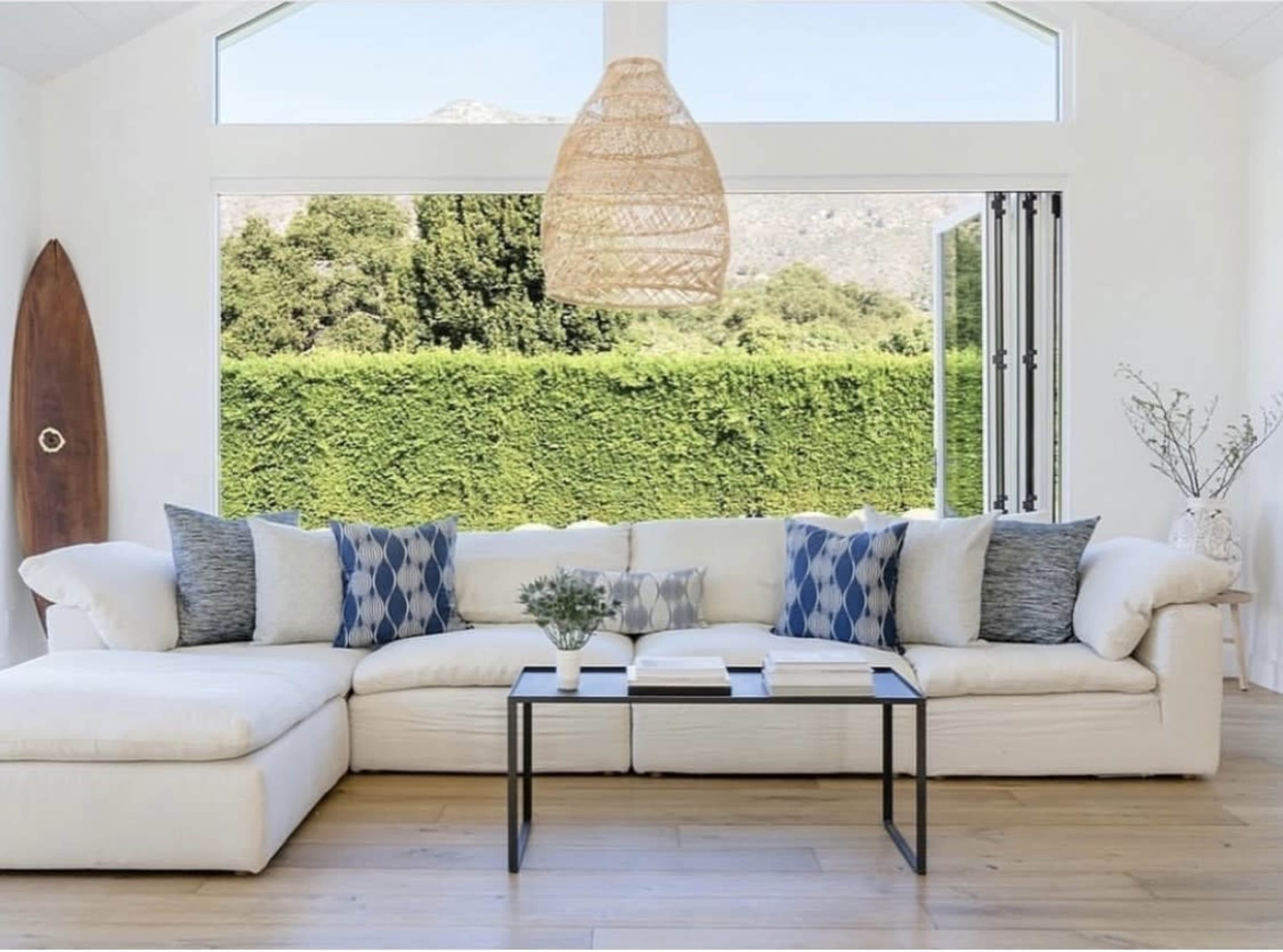 A spacious living room features a large white sectional sofa with decorative blue and gray pillows, a black coffee table, and a view of a green hedge through tall glass doors.