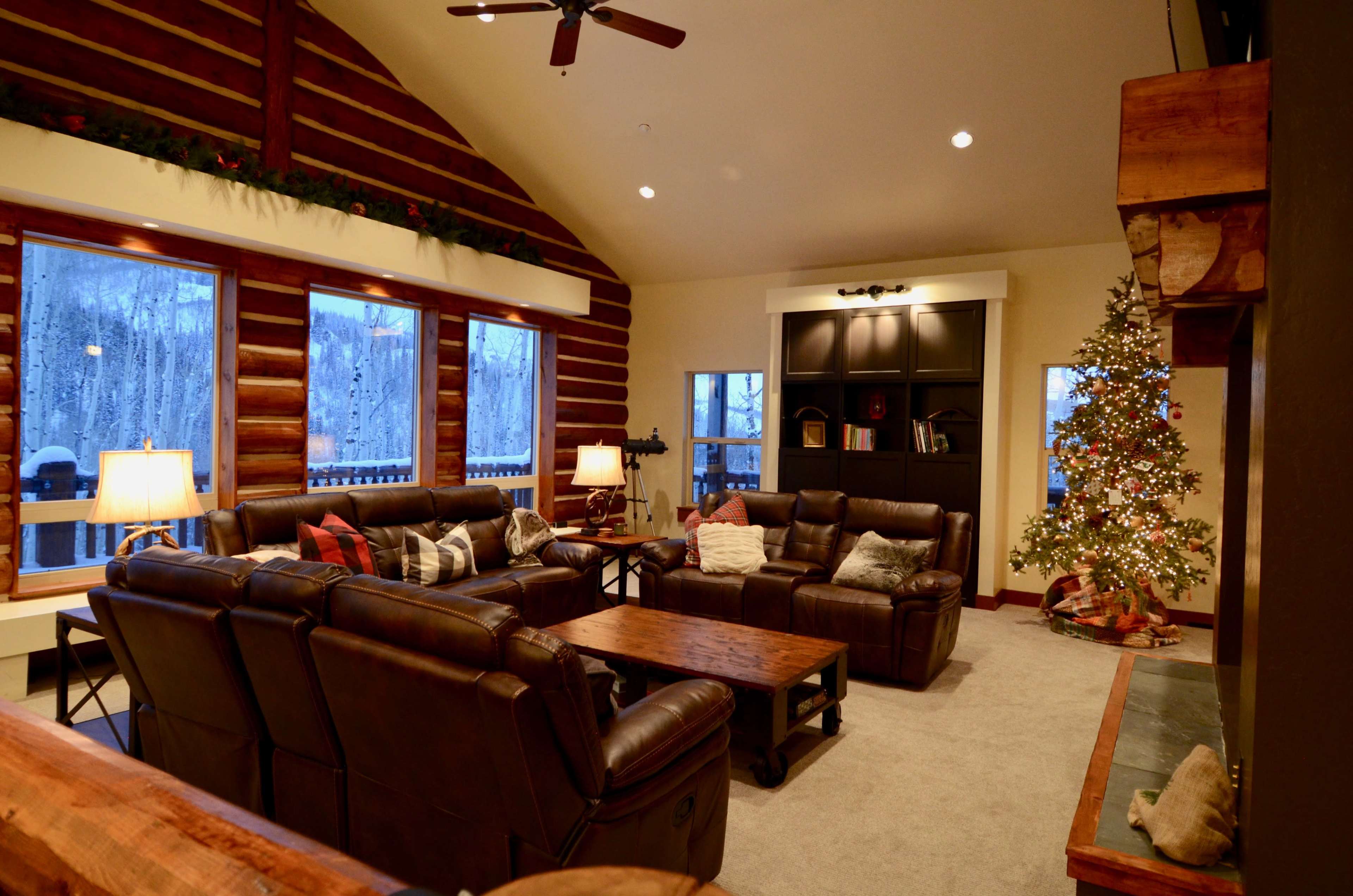 The cozy living room features leather sofas, a wooden coffee table, large windows showcasing a snowy landscape, and a decorated Christmas tree.