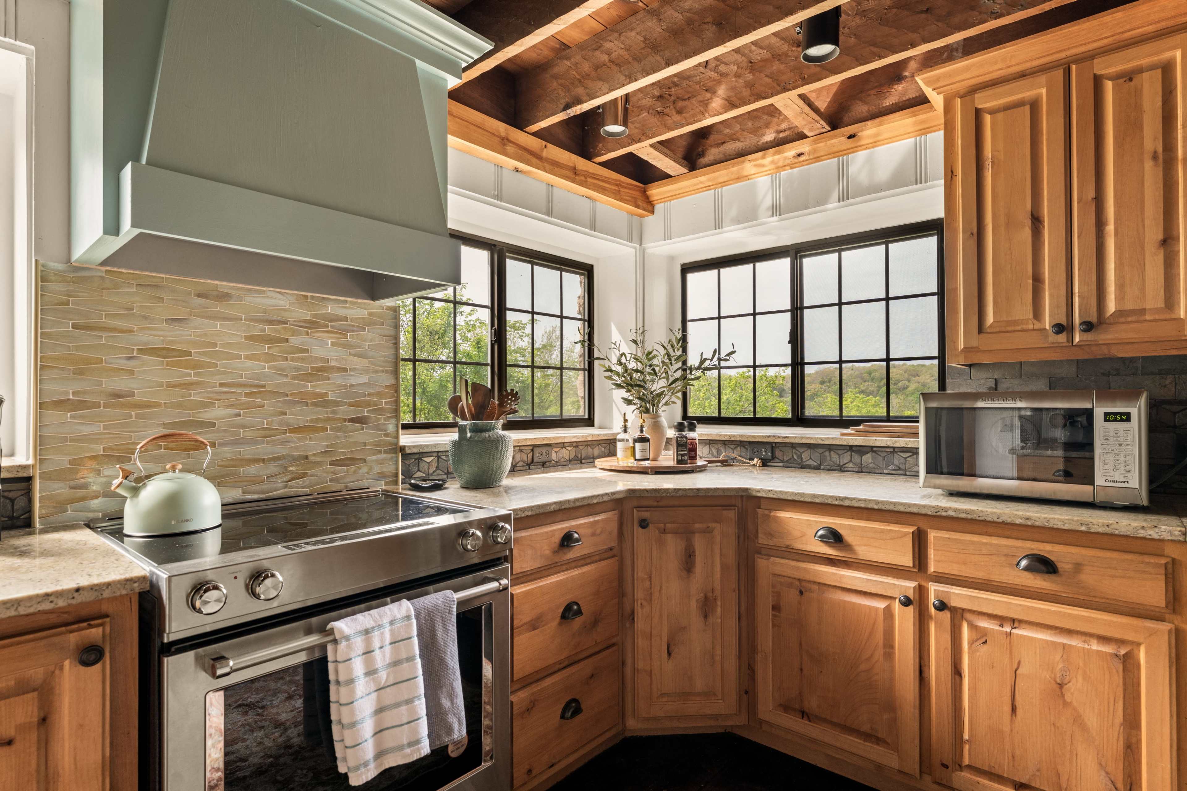The image shows a kitchen featuring wooden cabinets, a stainless steel stove with a light blue hood, and large windows allowing natural light to enter.