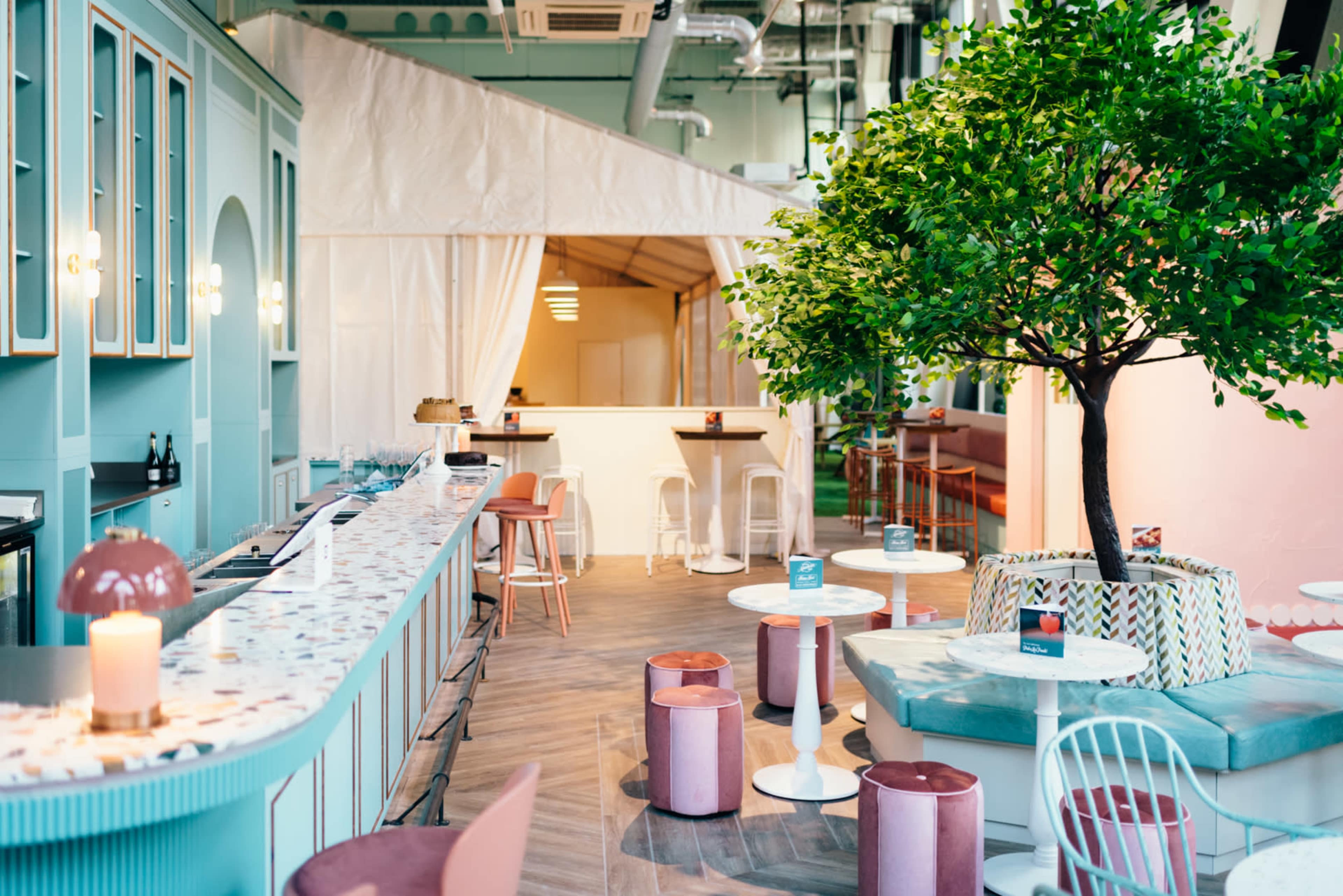 The image shows a brightly lit café interior featuring a pastel-colored bar, tables, and a decorative tree.
