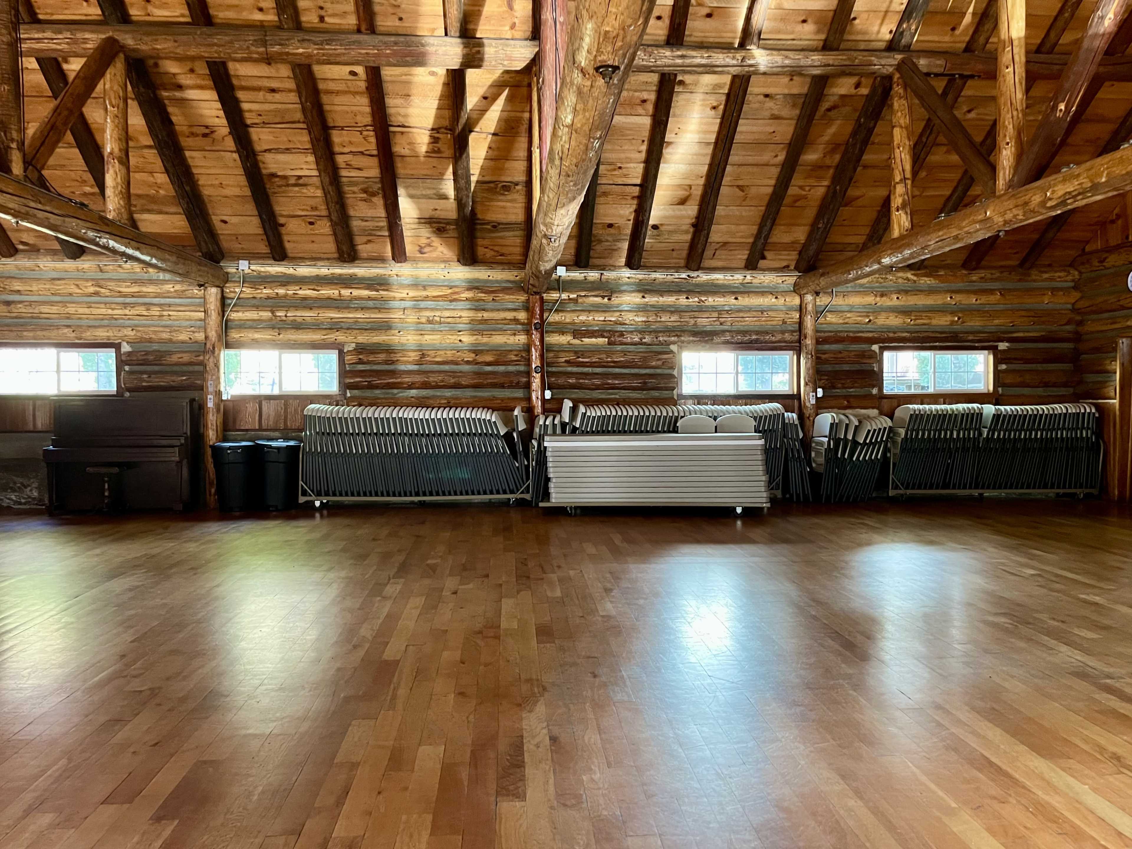 A spacious room with wooden floors and a high, exposed beam ceiling, featuring stacked folding tables against one wall.