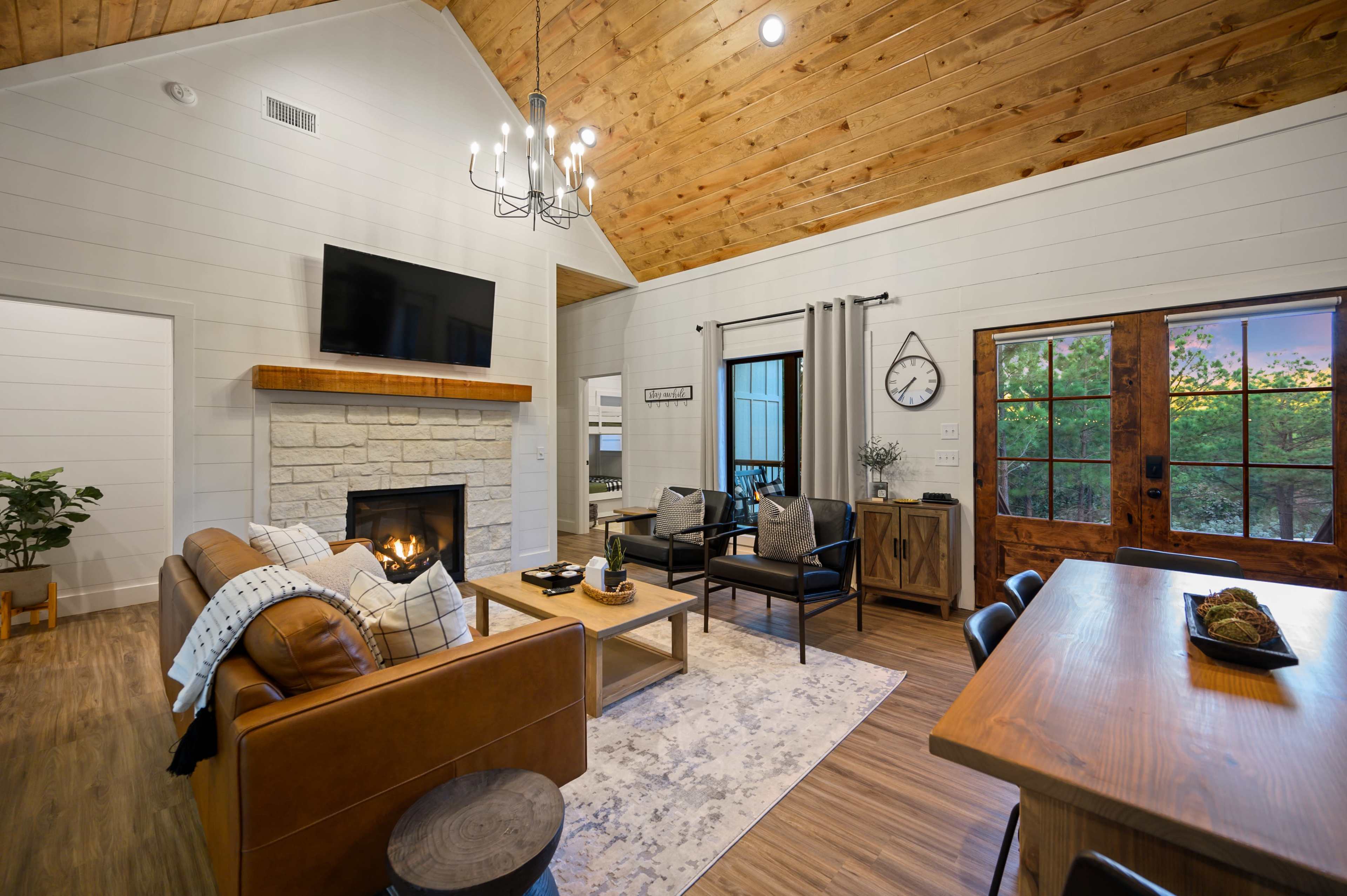 A cozy living space features a stone fireplace, a large flat-screen TV, and a dining area with wooden furniture beneath a vaulted ceiling.