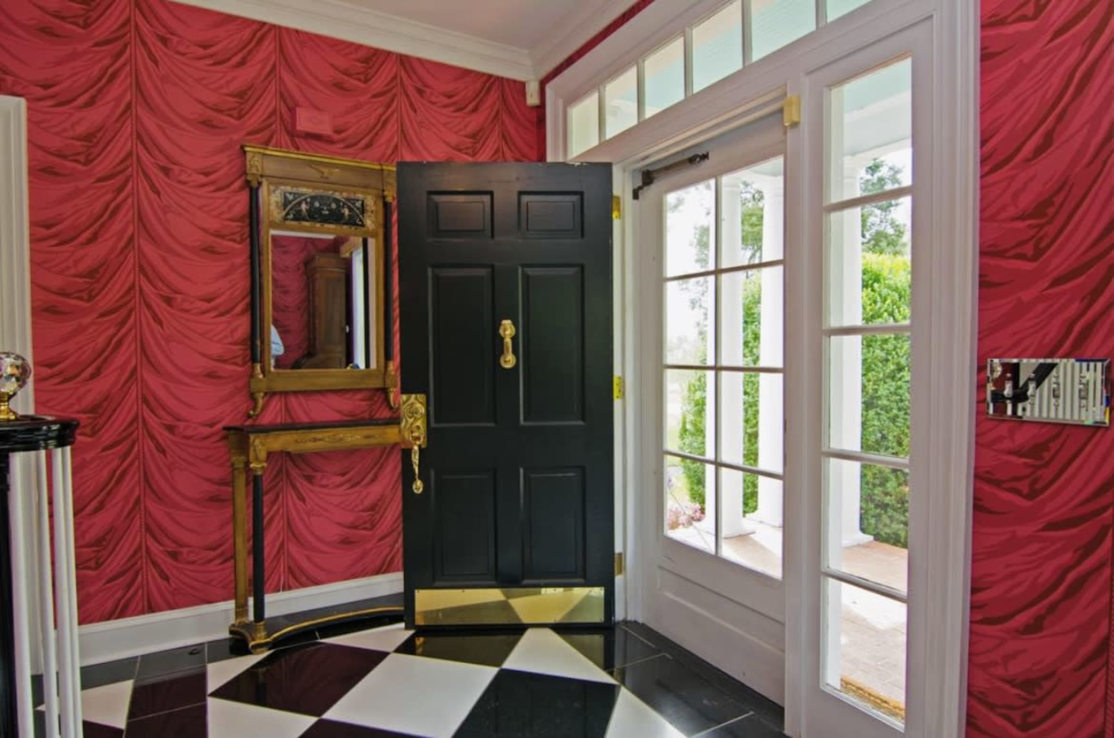 The entryway features a black front door with a gold handle, surrounded by pink textured wallpaper and a mirror above a wooden console table, with checkered flooring.