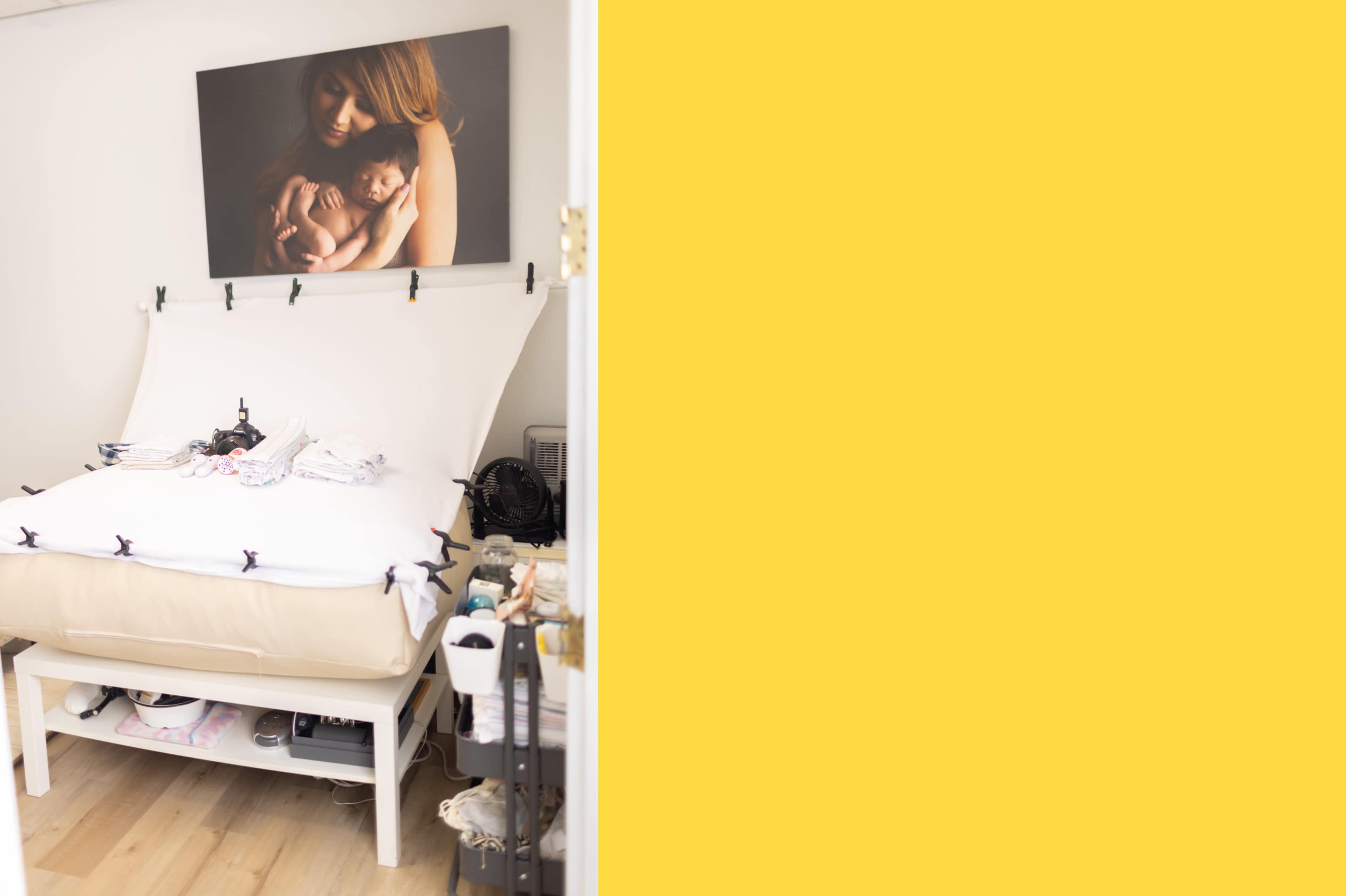 The image shows a bedroom with a bed covered by a white blanket, a large framed photograph of a mother and child on the wall, and a light yellow section on the right.