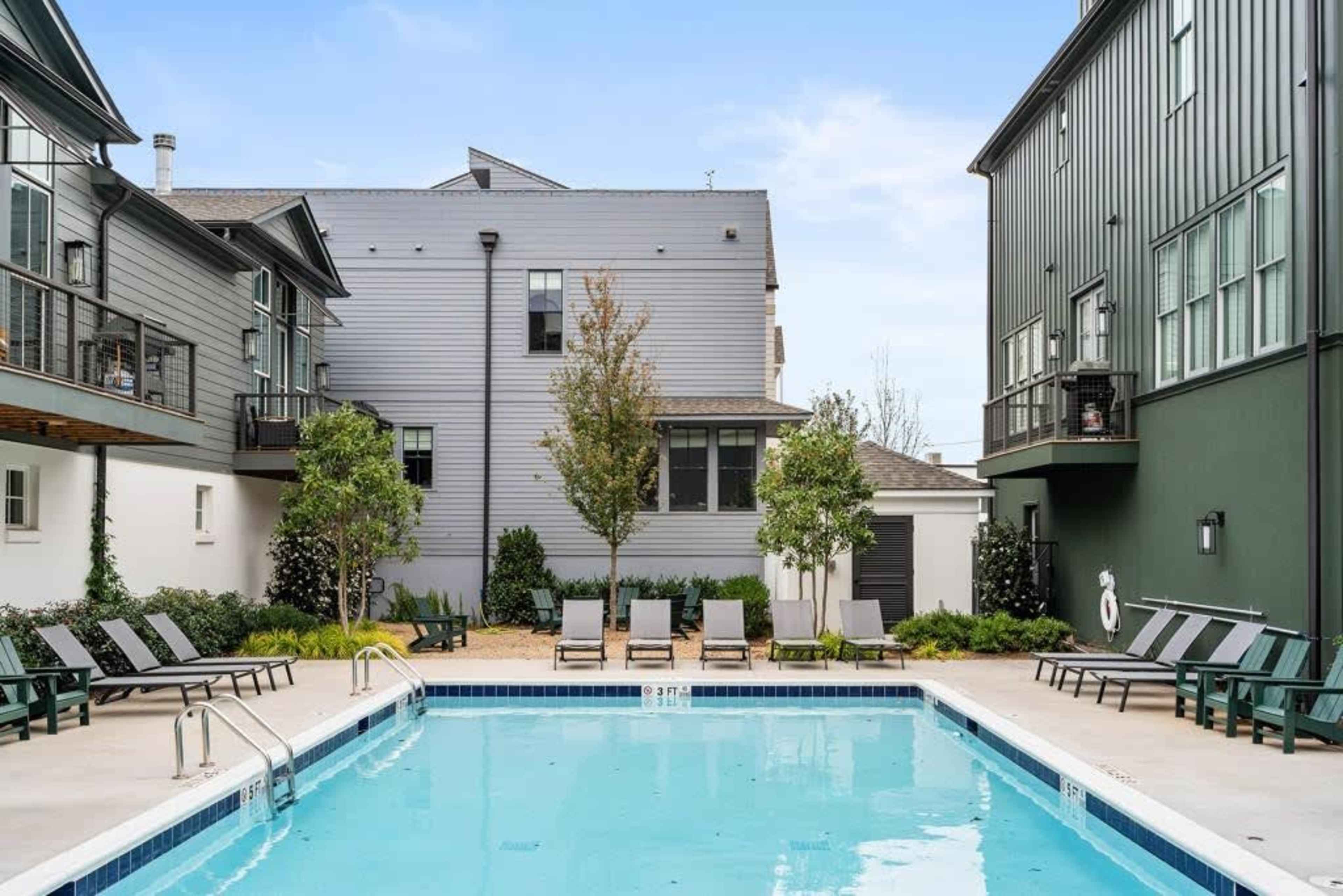 A swimming pool is surrounded by chaise lounges and small trees, with two multi-story buildings in the background.