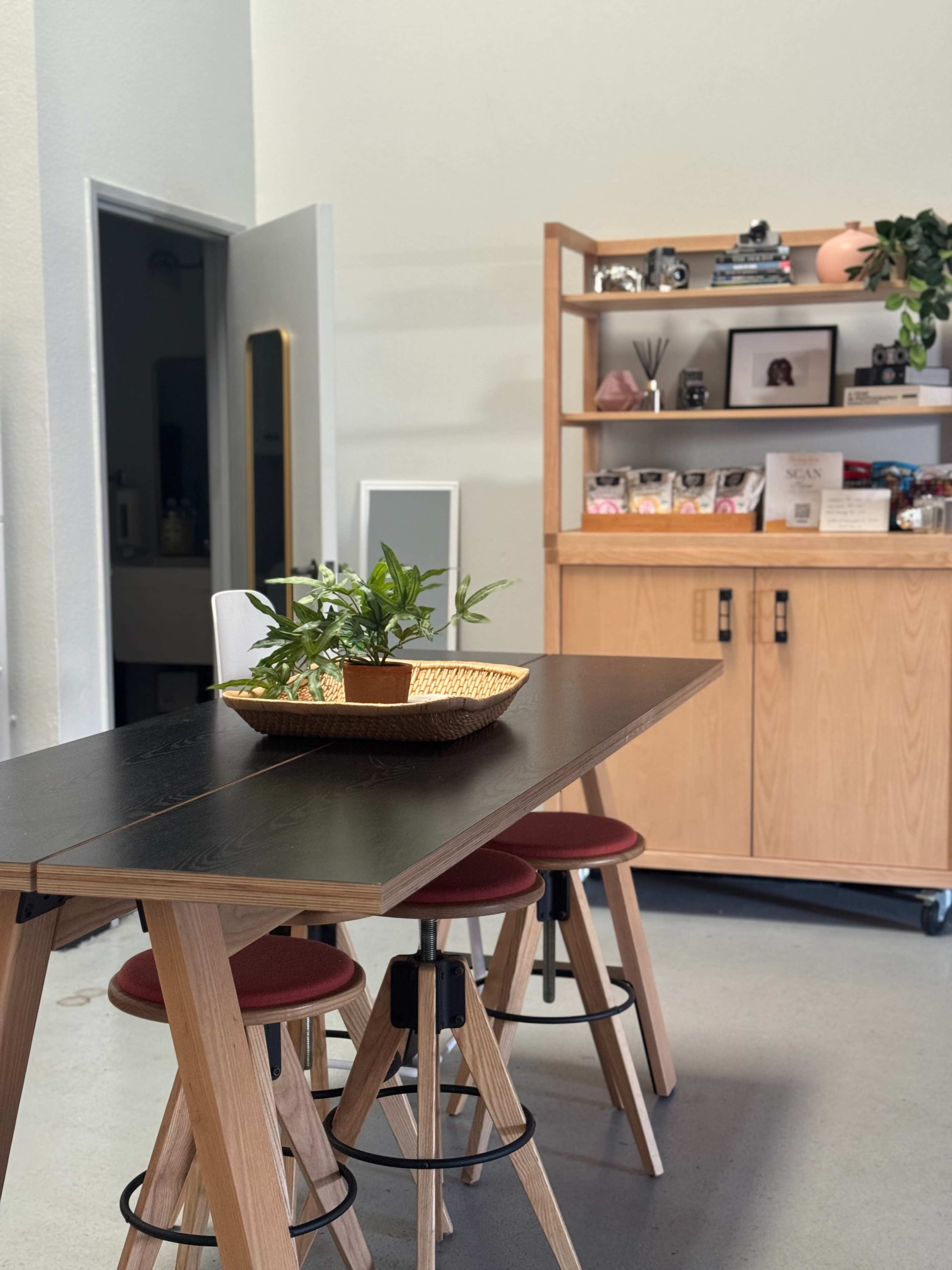 The image shows a modern kitchen area with a wooden table and stools, a plant centerpiece, and a cabinet filled with decorative items and books.