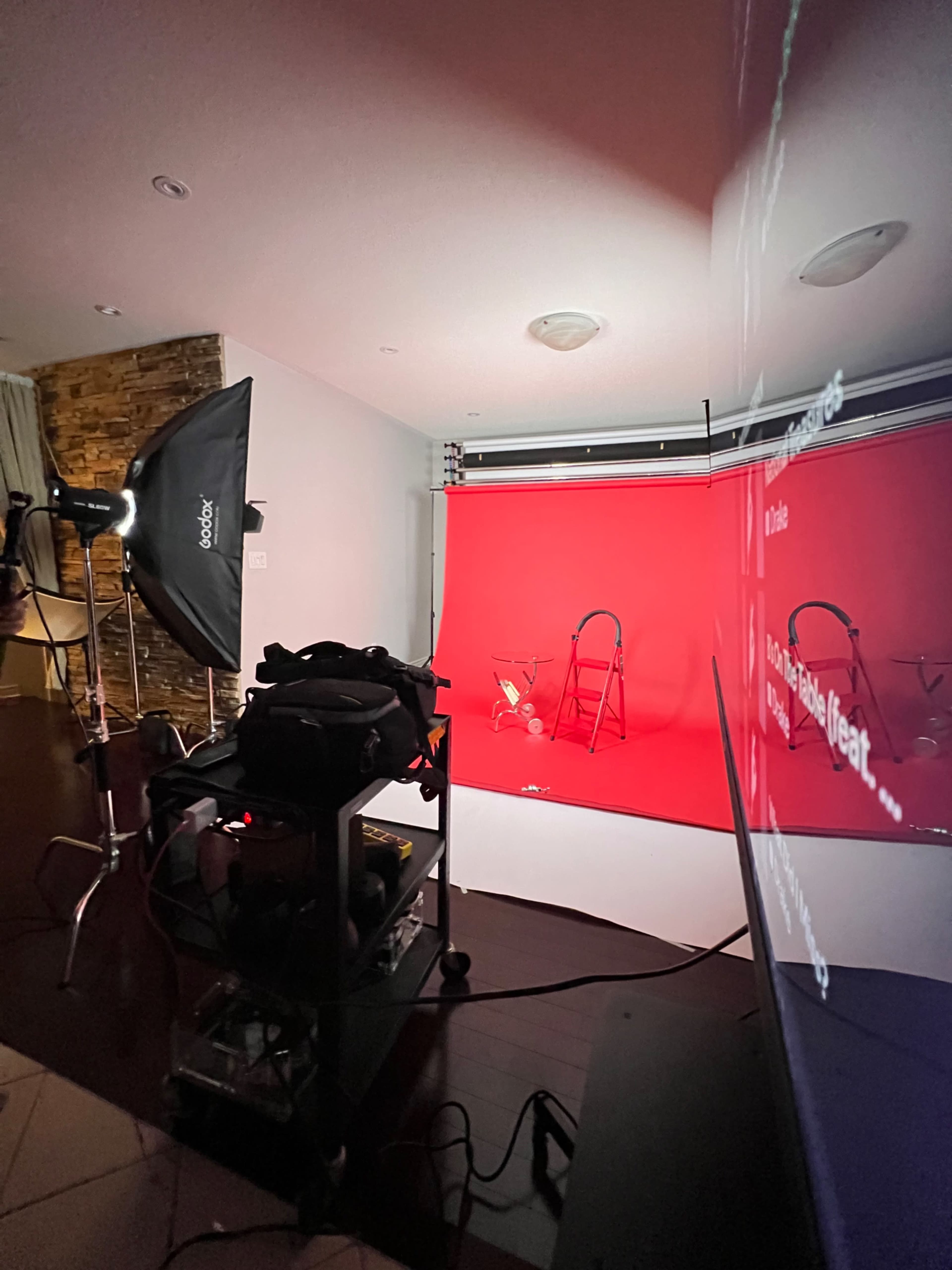 The image shows a photography setup in a room featuring a red backdrop, a light source on a stand, and a black cart with equipment nearby.