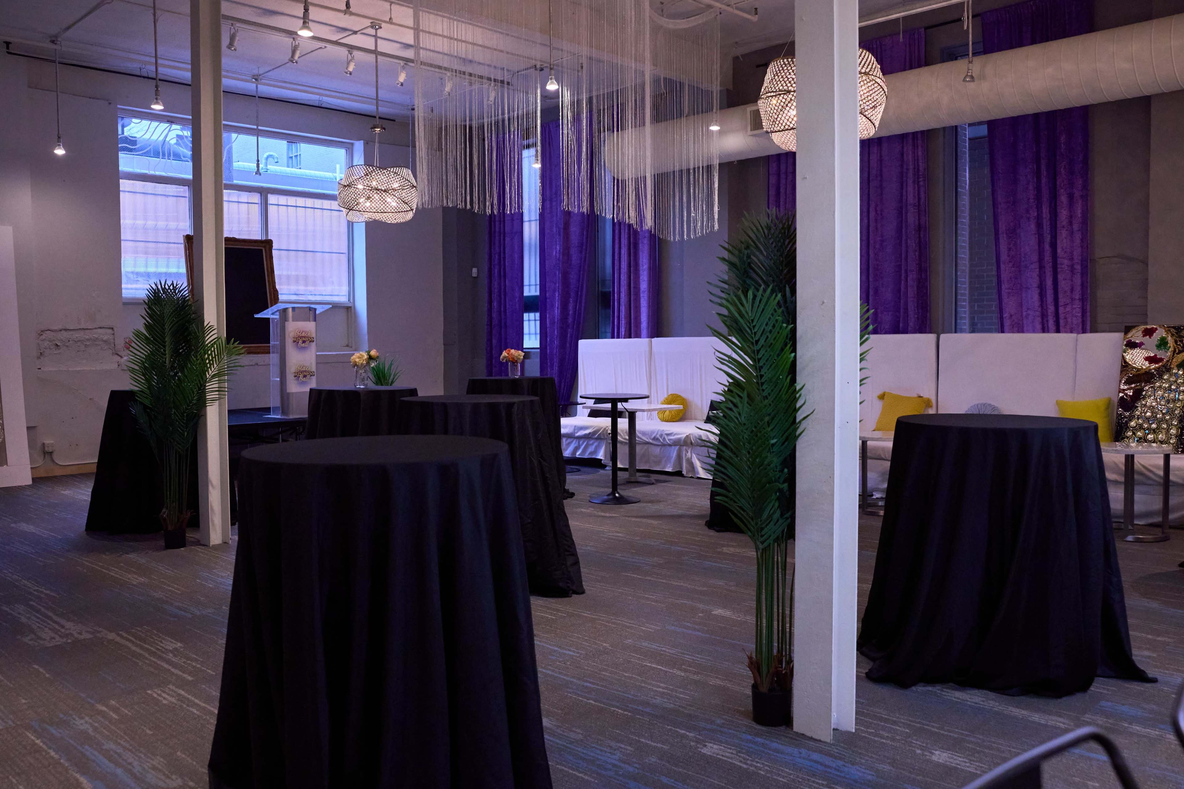 A spacious event room features several tall tables with black tablecloths, surrounded by minimalist decor and soft lighting.