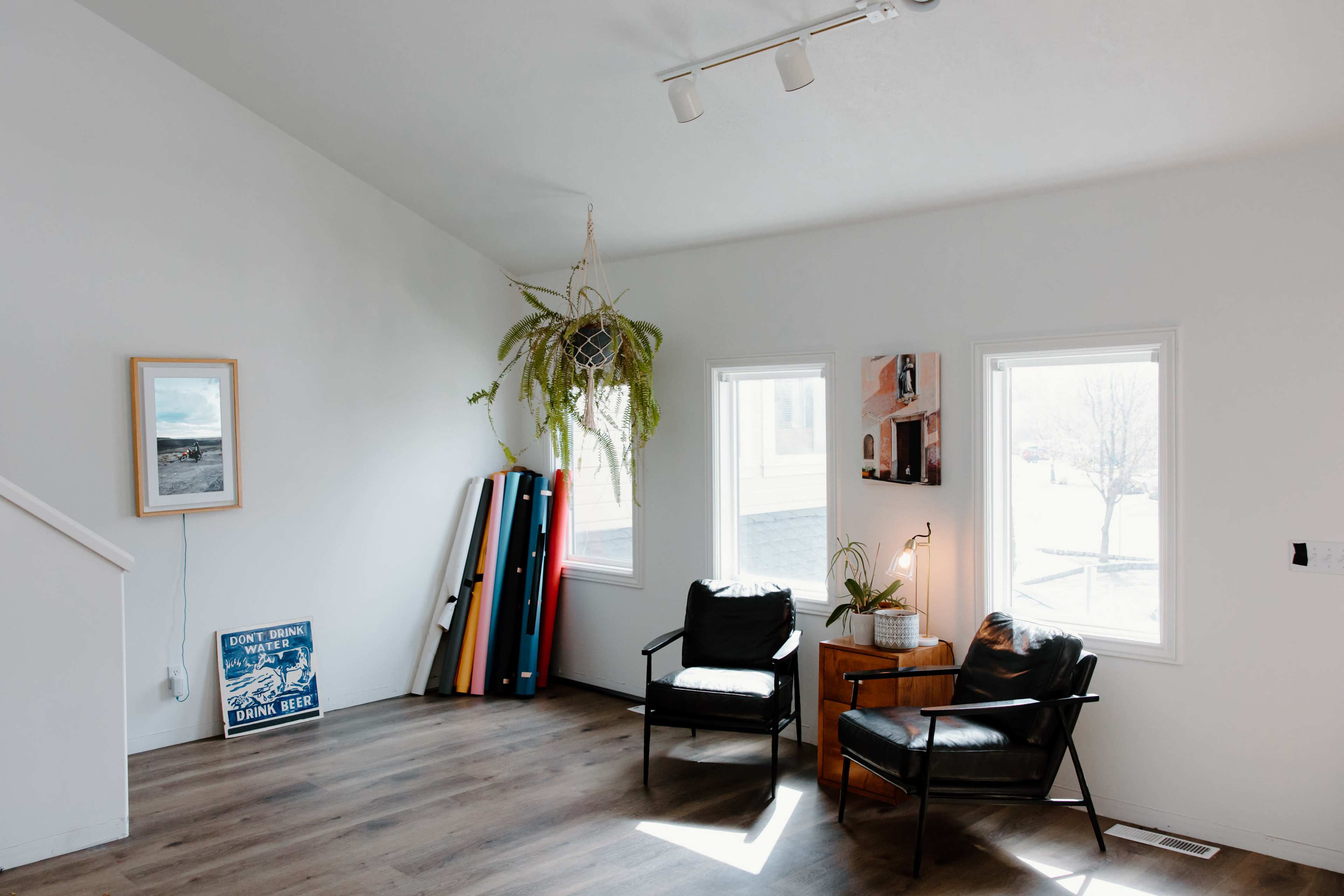 A bright room features two black chairs, a wooden side table, large windows, and colorful boards leaning against the wall.