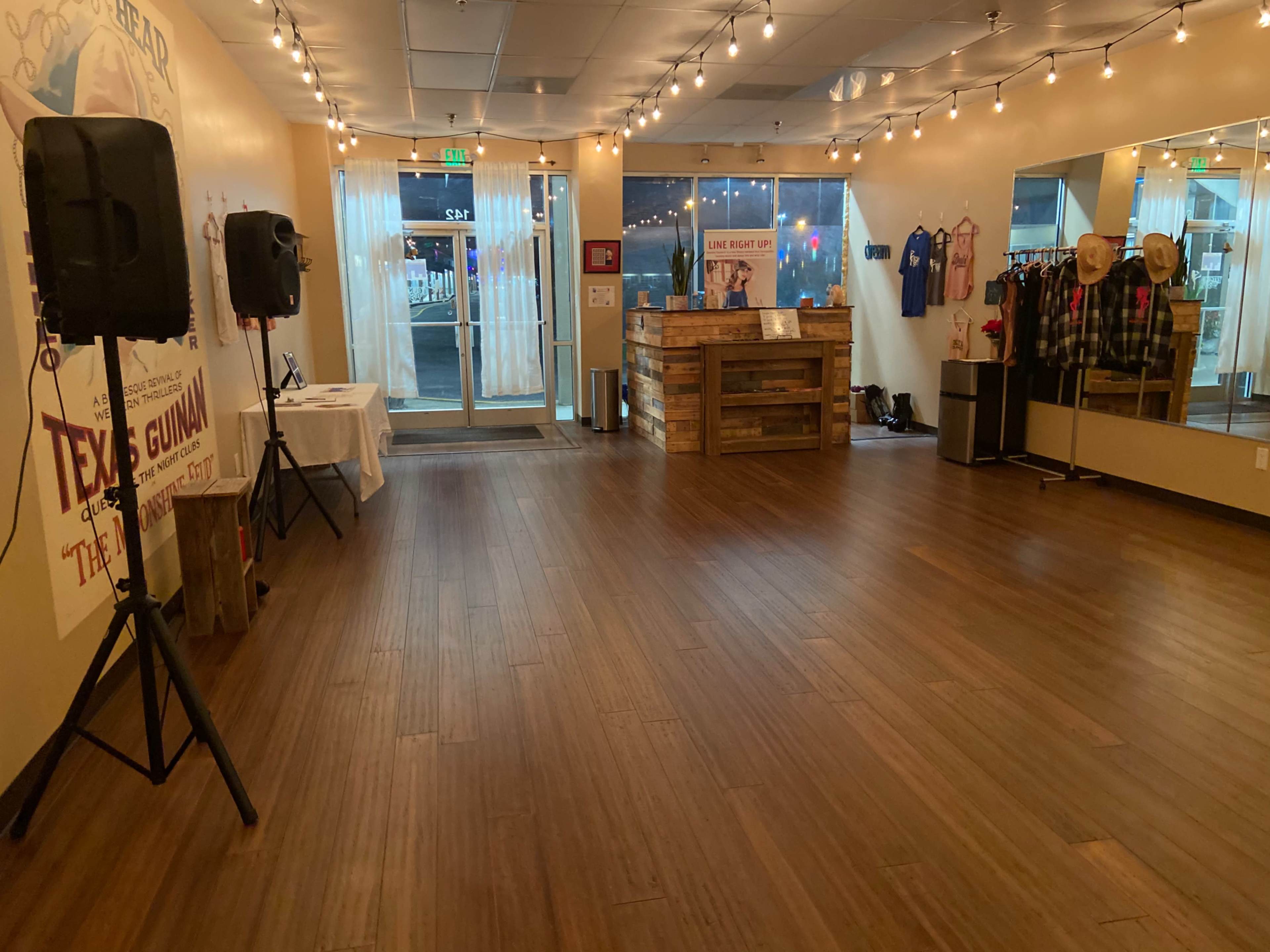 An empty dance studio with wooden flooring, speakers on stands, and a reception area featuring a wooden counter and mirrored walls.