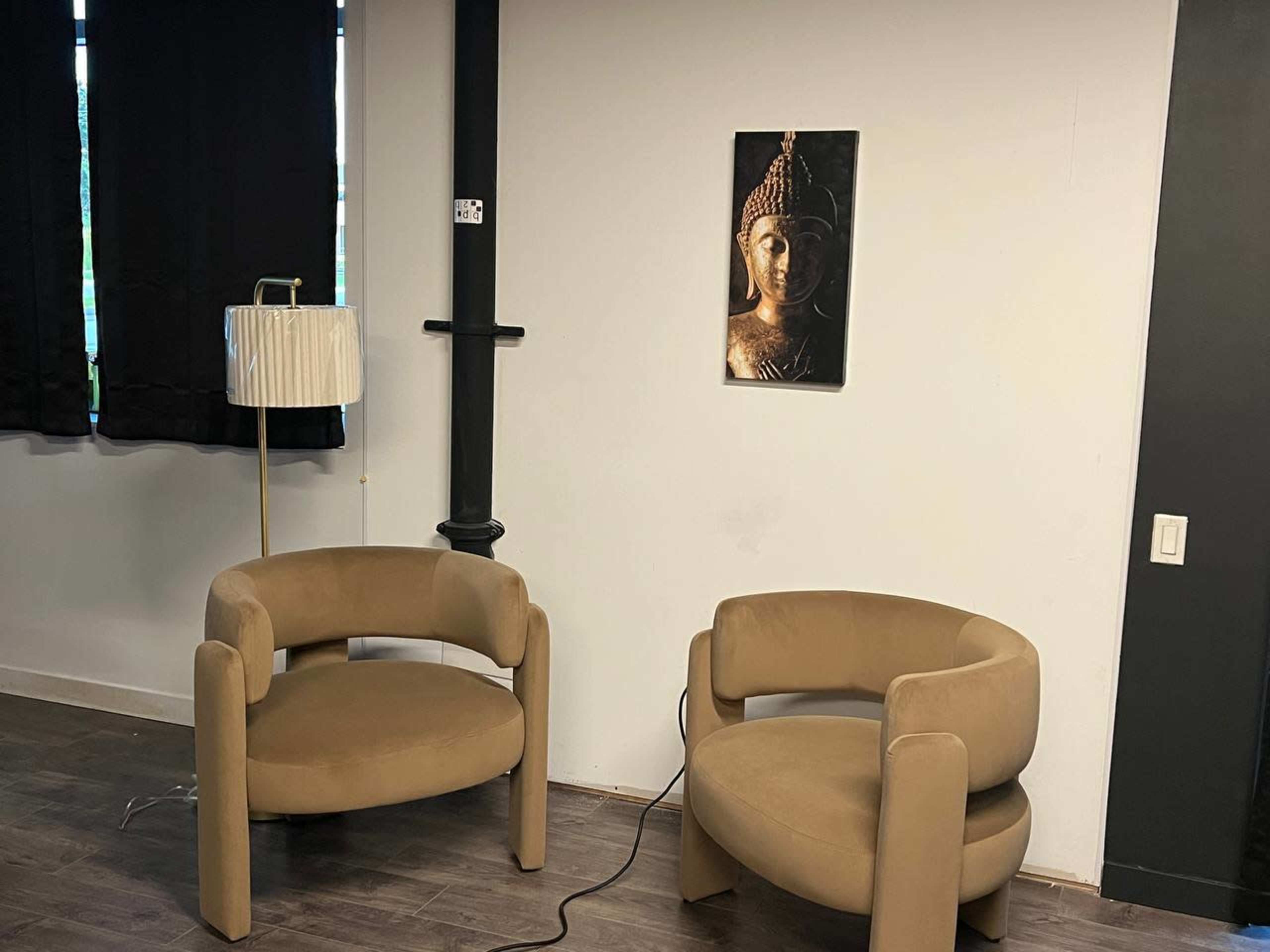 The image shows two beige, modern armchairs facing each other in a room with dark curtains and a wall-mounted artwork of a Buddha face.