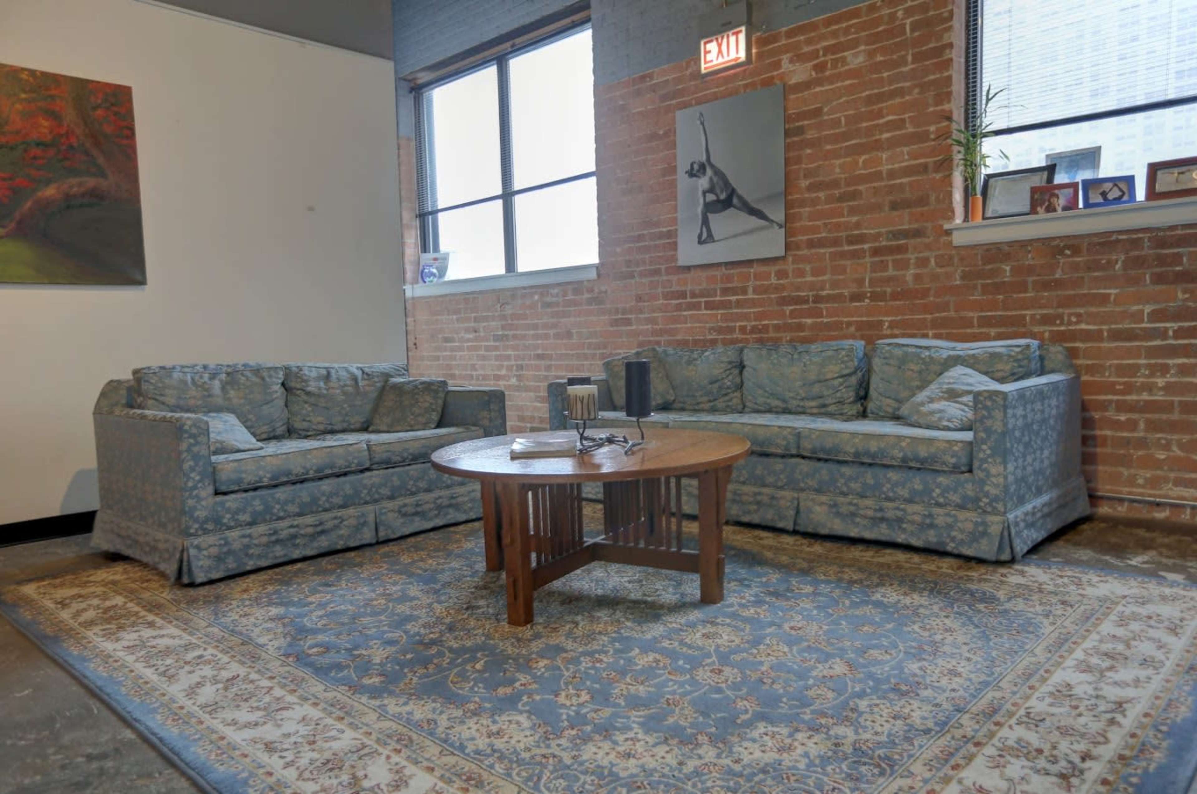 A cozy living area with two patterned sofas facing a wooden coffee table on a decorative rug, against a backdrop of exposed brick walls and large windows.