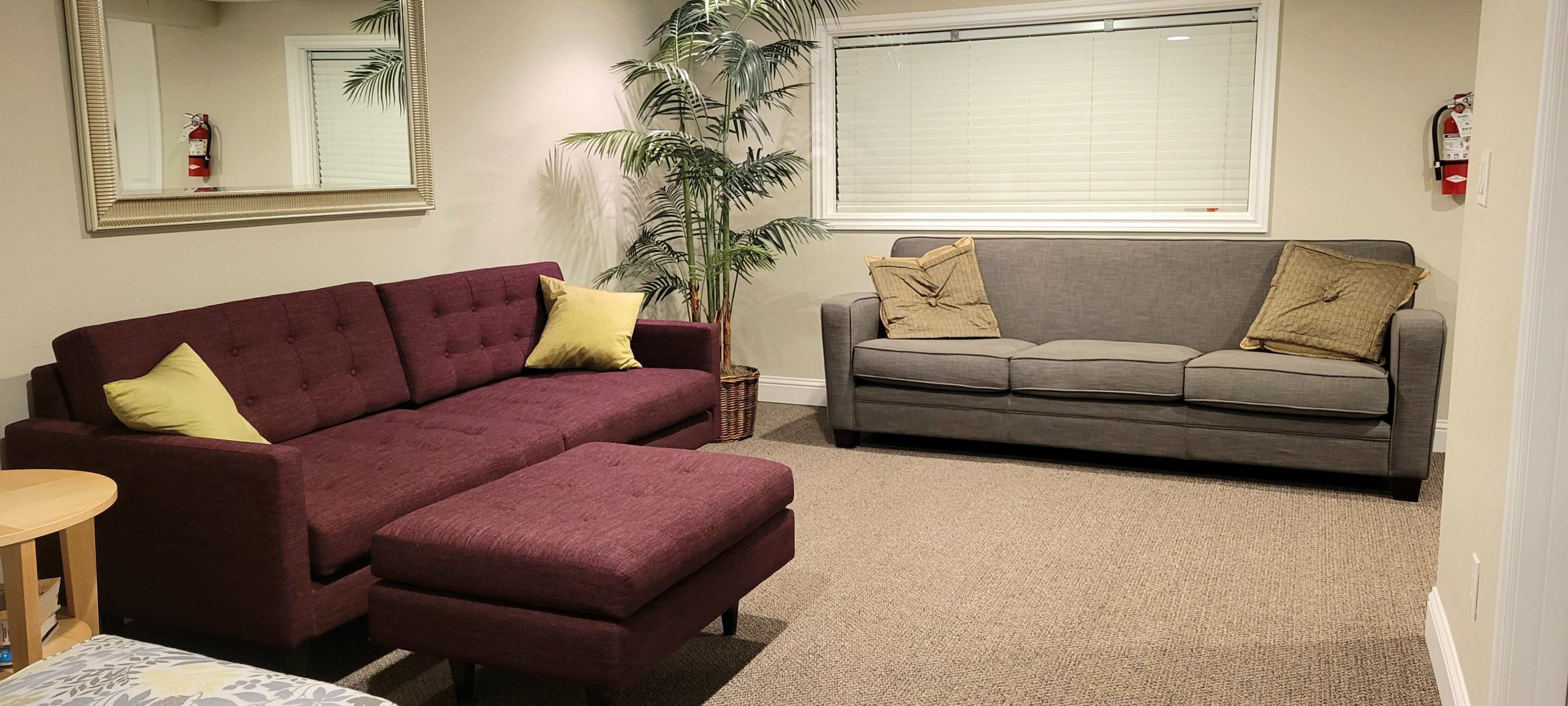 A room with a purple sectional sofa, an ottoman, and a gray couch, along with a plant and a mirror on the wall.