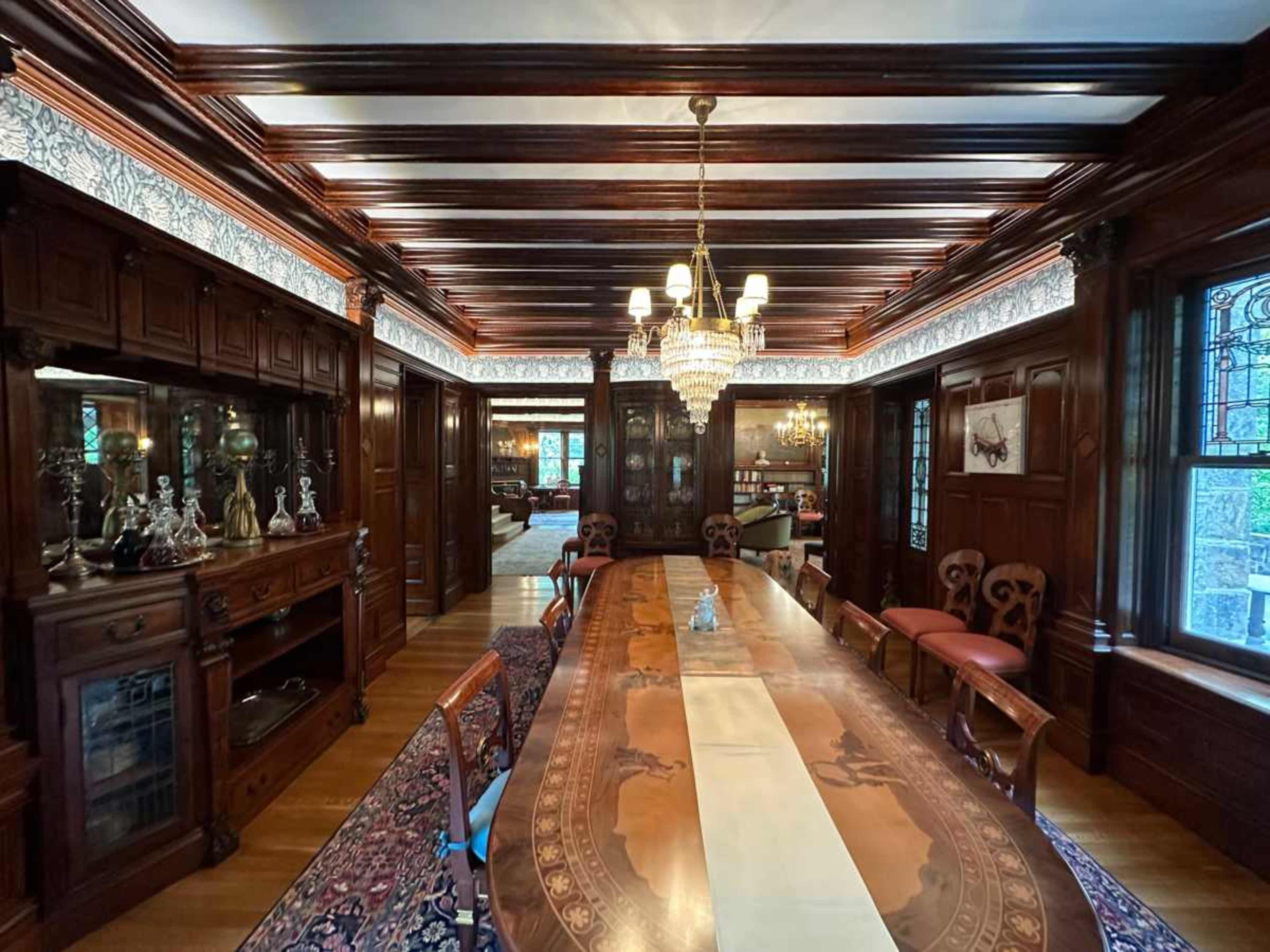The image shows an elegantly furnished dining room featuring a long wooden table with a decorative inlay, surrounded by high-backed chairs, and illuminated by a chandelier.