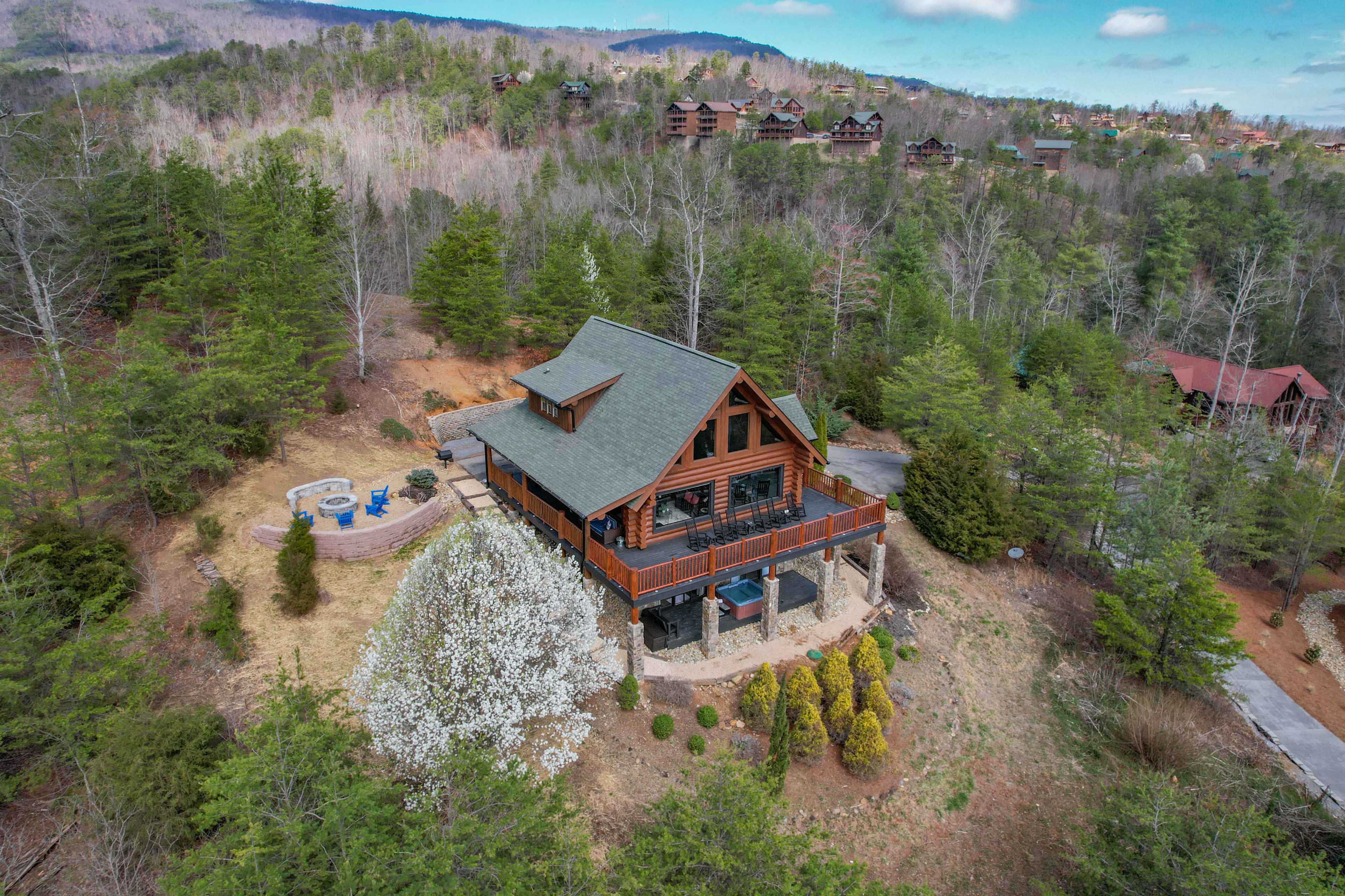 A large, wooden cabin is nestled amongst trees and hills, featuring a deck and outdoor seating area with a fire pit.