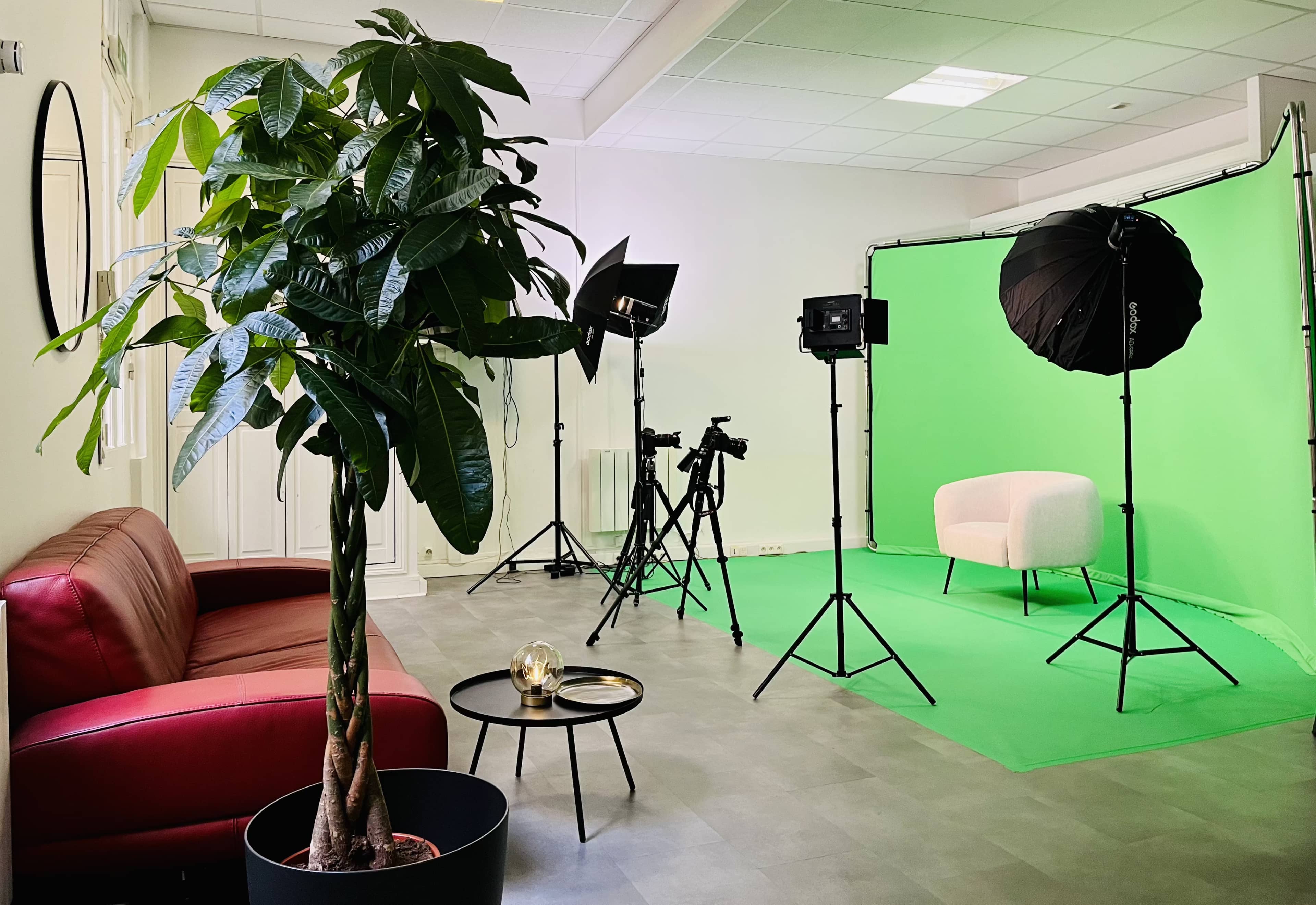 A studio setup features a green backdrop, lighting equipment, cameras on tripods, a white chair, and a gray floor, with a potted plant and a brown sofa in the foreground.