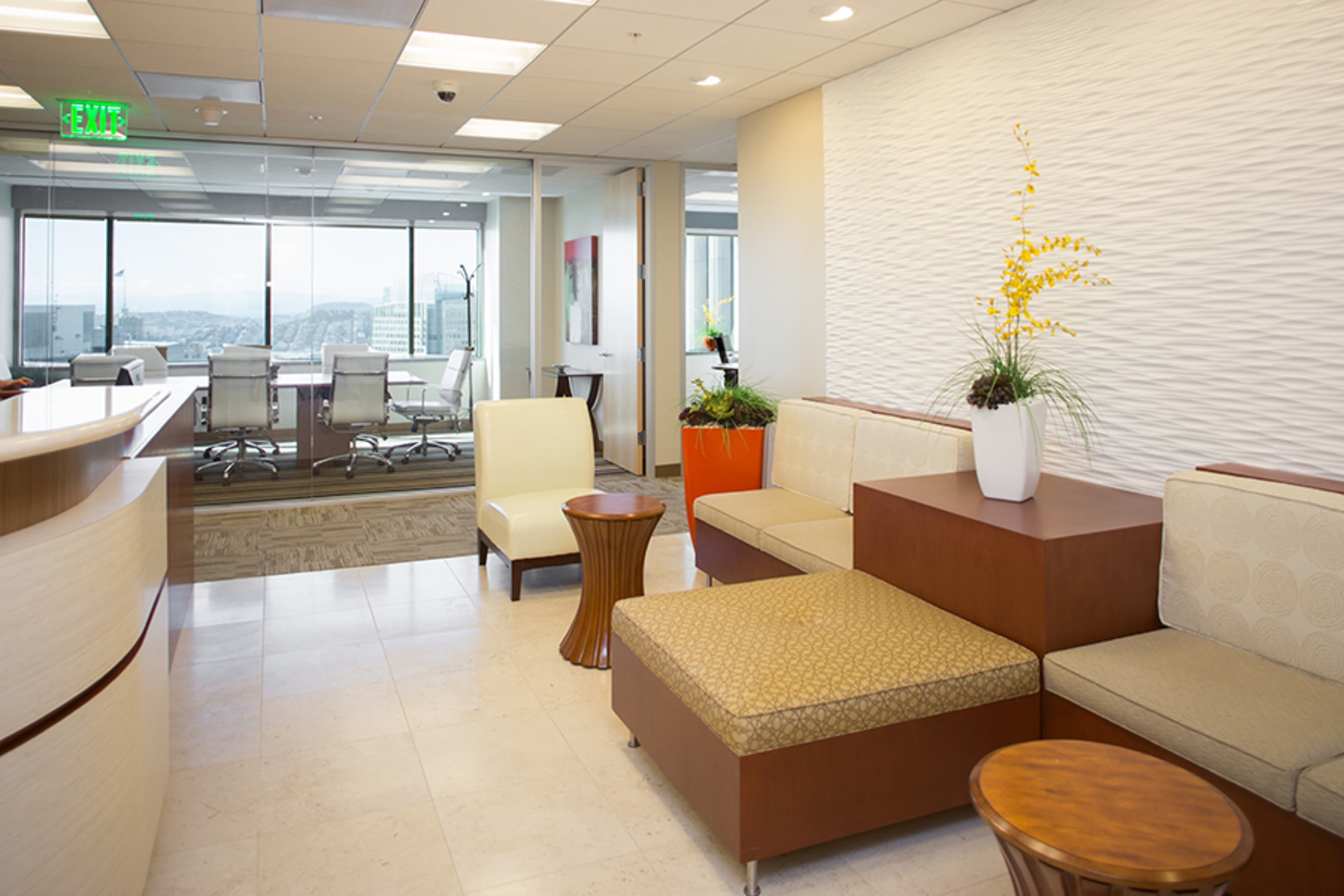 A modern office waiting area with seating, a small table, and a view of a glass-paneled conference room in the background.