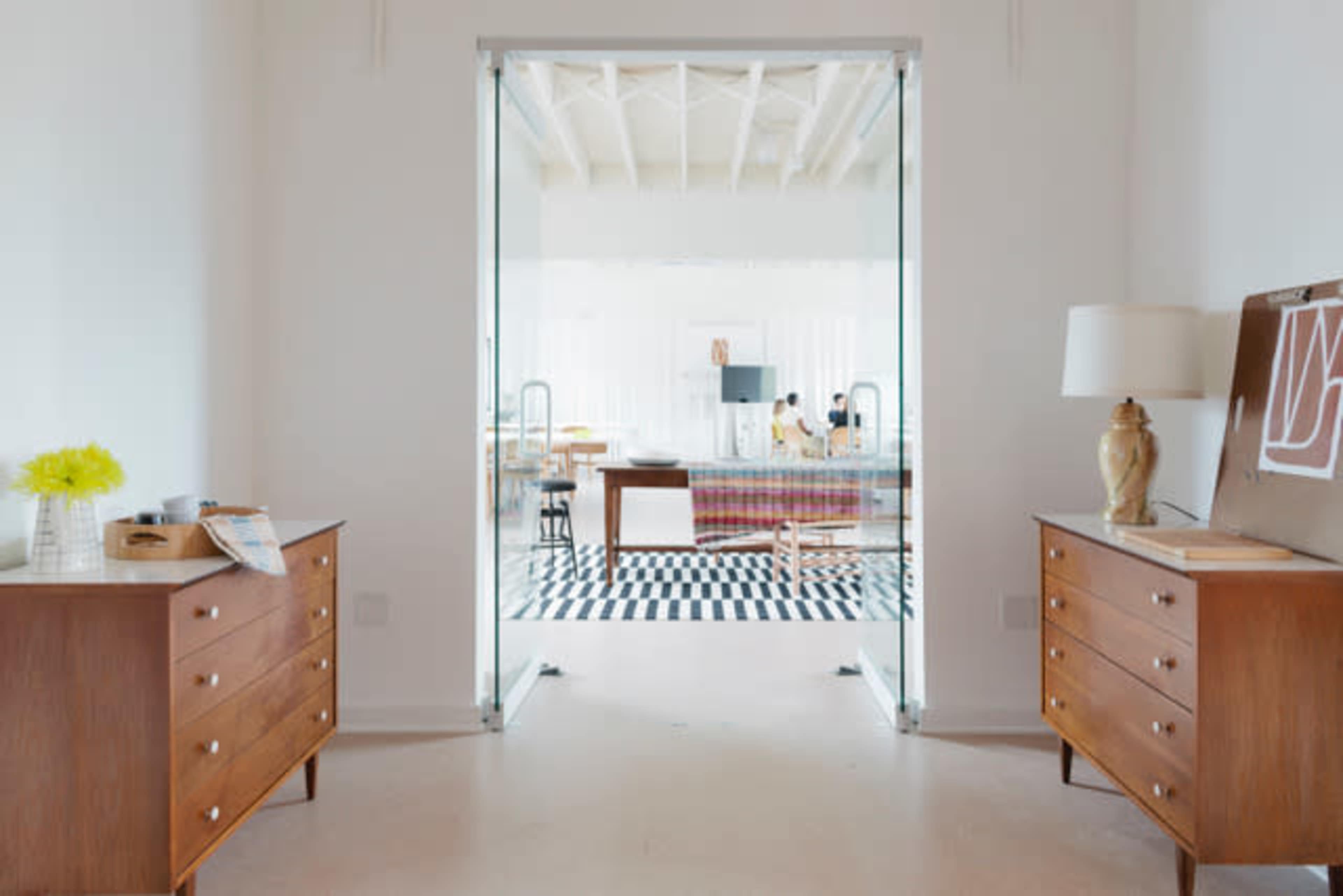 A modern interior features a glass doorway leading into a spacious room with a checkered rug, wooden furniture, and a decorative lamp.