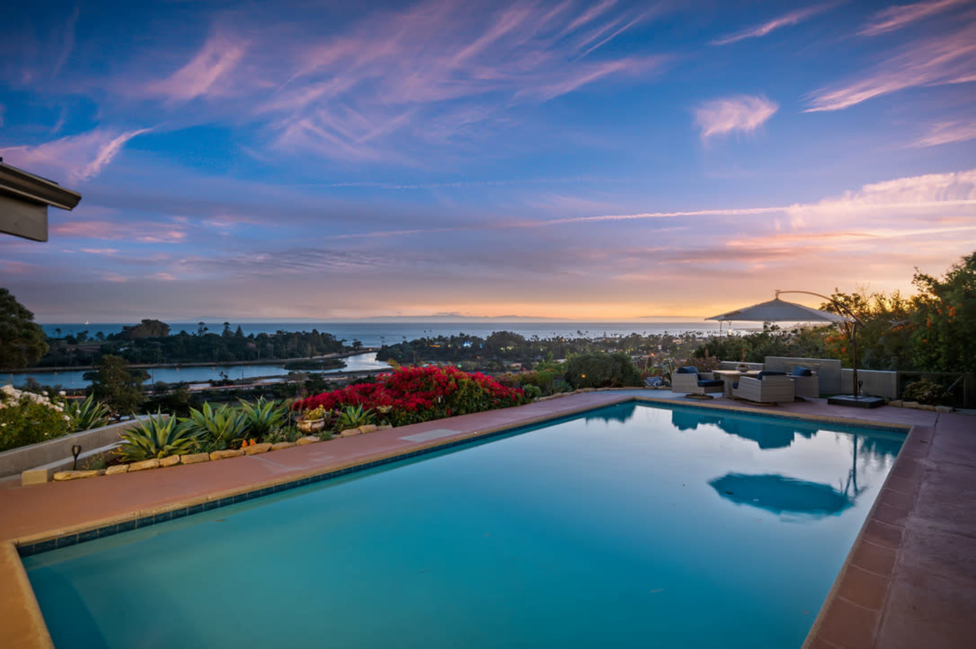 A swimming pool reflects the colorful sunset over a coastal landscape with lush greenery and flowering plants.