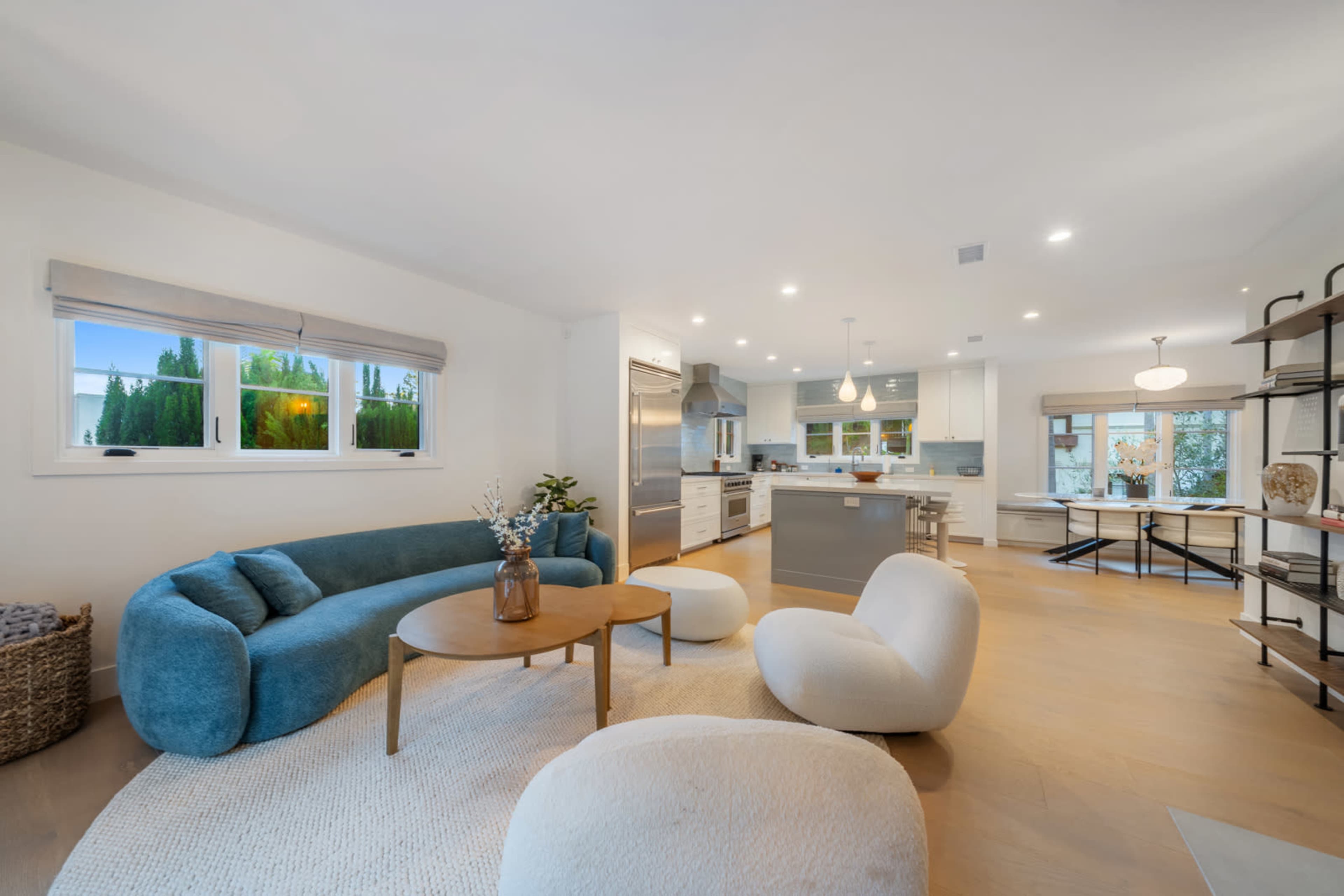 The image features a modern living space with a blue curved sofa, two white lounge chairs, a round coffee table, and a kitchen area visible in the background, illuminated by bright, natural light.