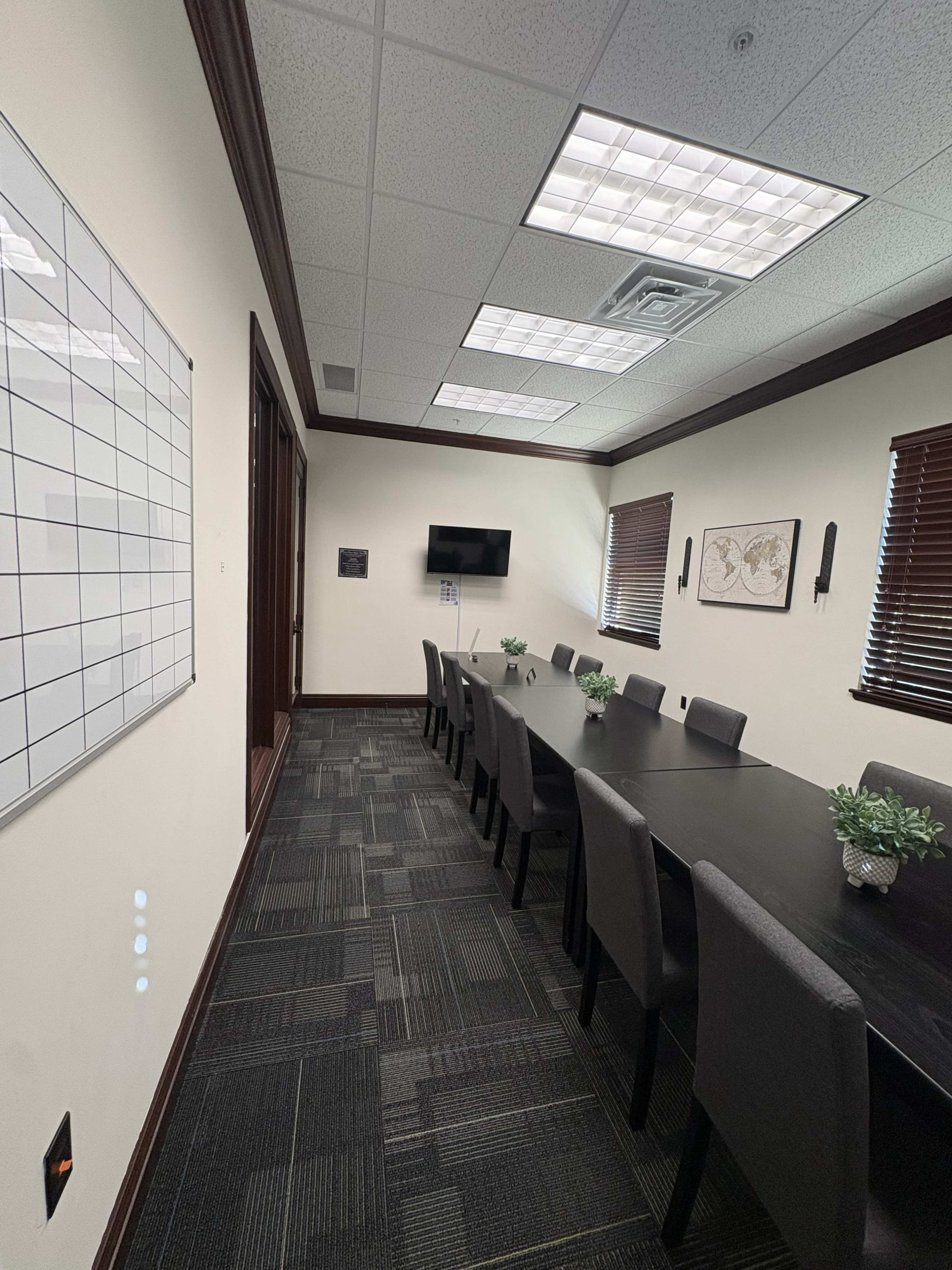 A long conference table with chairs is placed in a well-lit meeting room featuring a whiteboard, a television, and decorative plants.