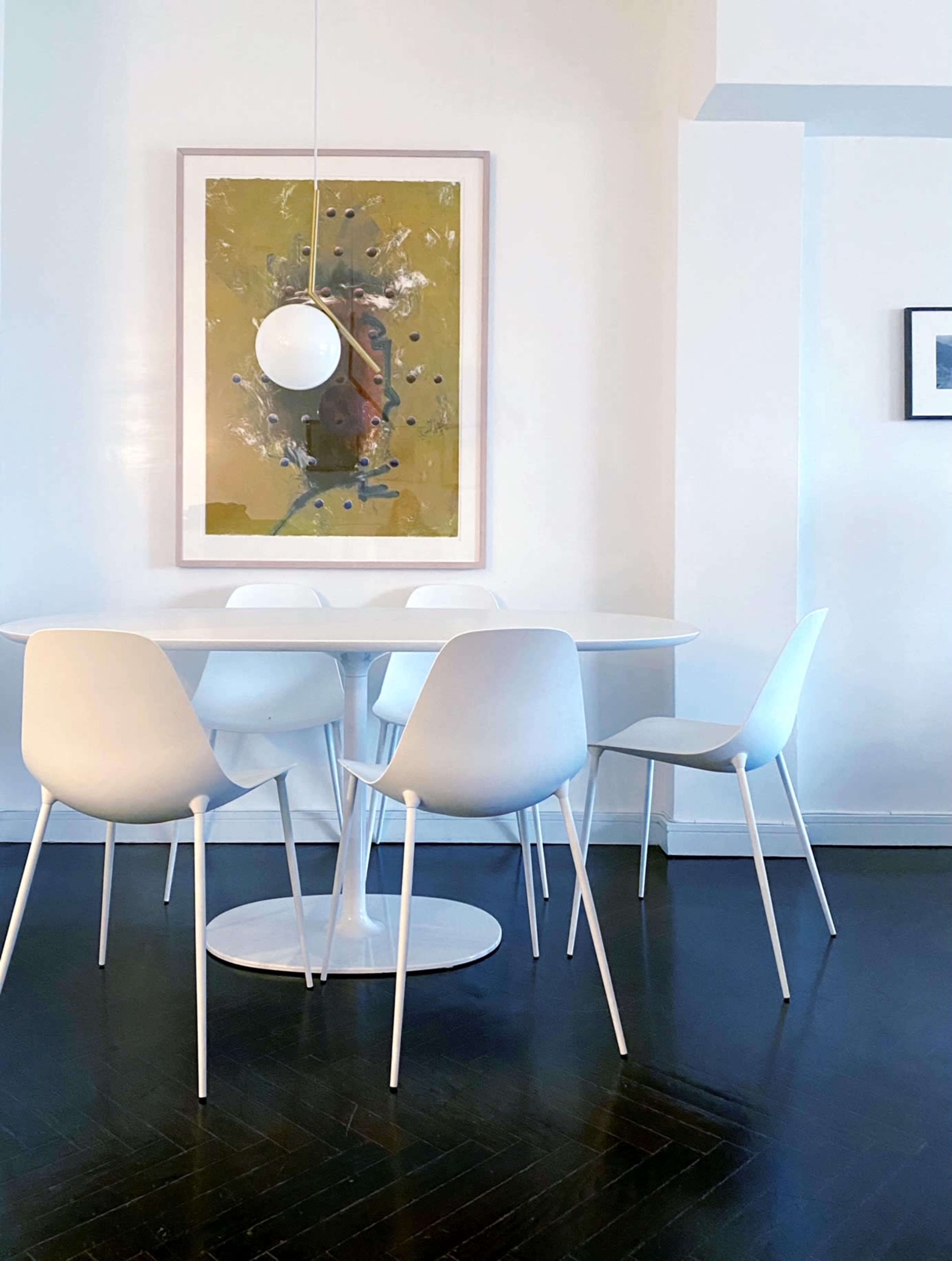 A modern dining area features a round white table surrounded by four white chairs, with a large piece of abstract art on the wall and a pendant light hanging above.