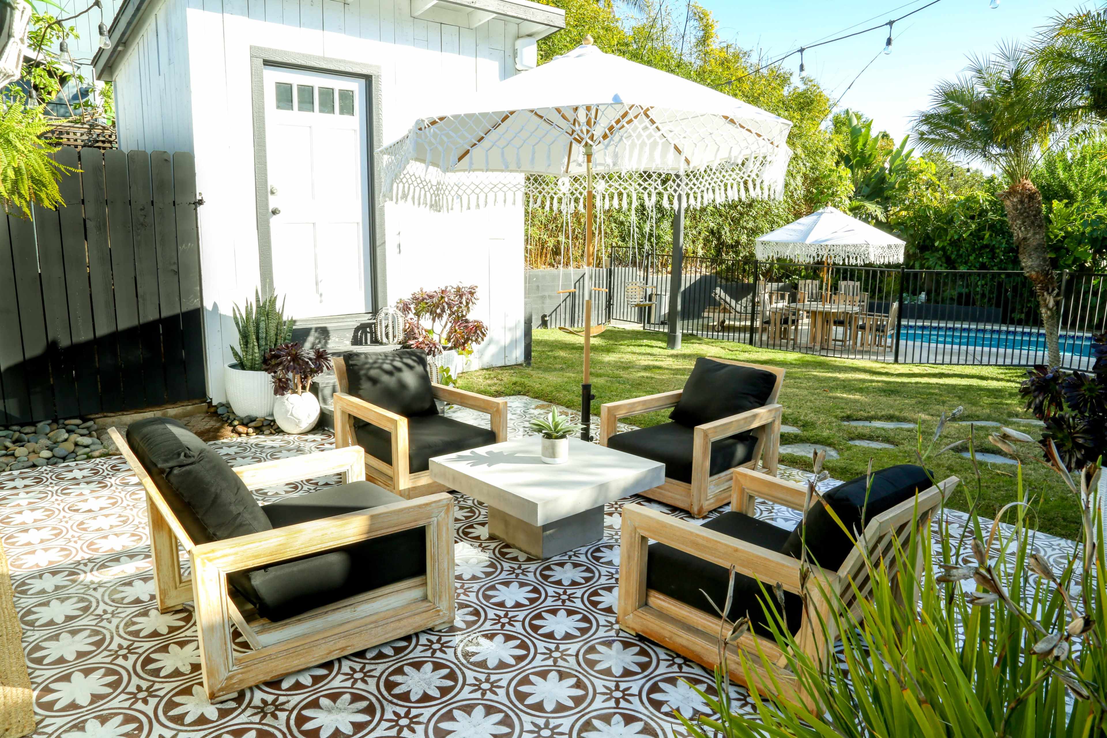 A modern outdoor seating area with black cushioned chairs and a white table, surrounded by greenery and a patterned tiled floor.